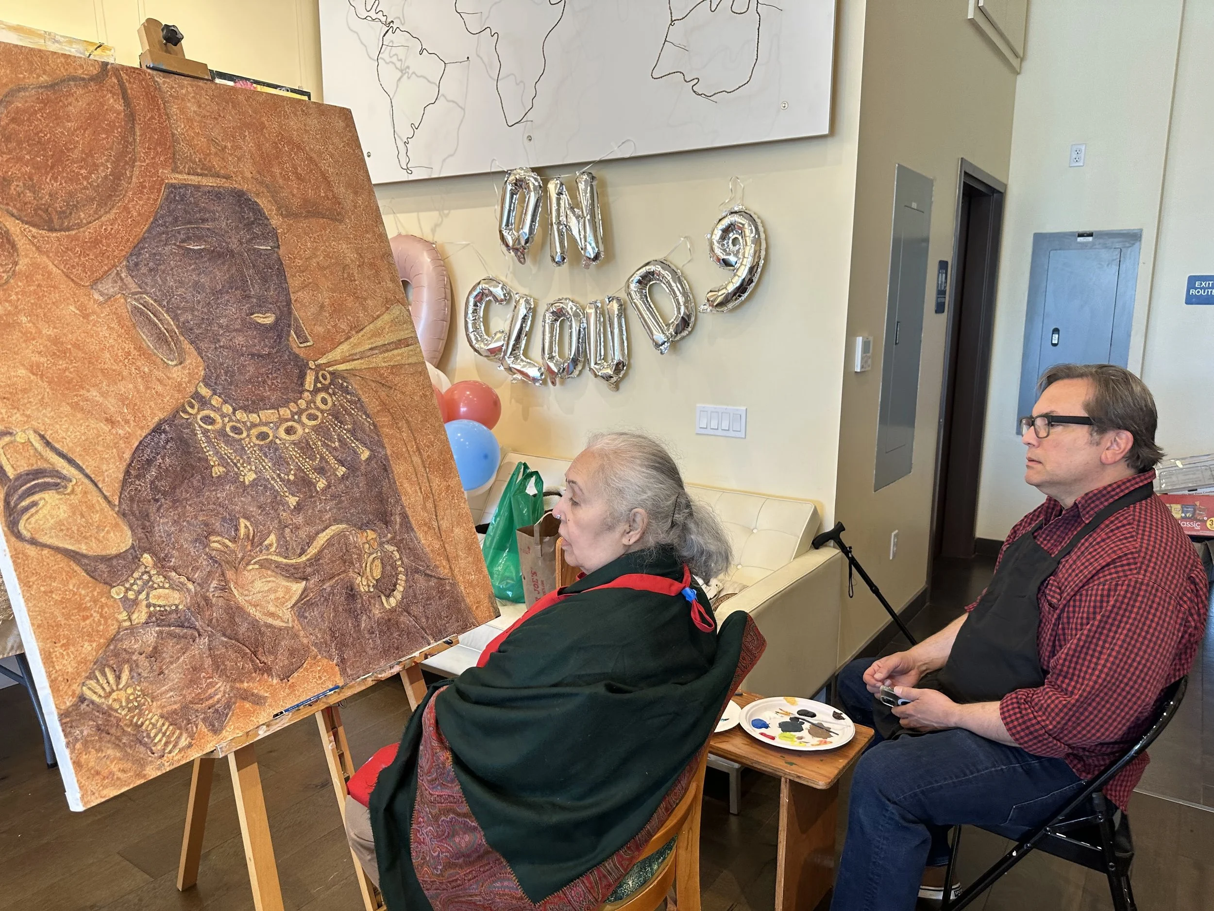 An elderly woman and a man sit and observe an artwork of a woman with dark skin wearing elaborate jewelry, being painted with warm tones. There are balloons and balloons with letters on the wall behind them.