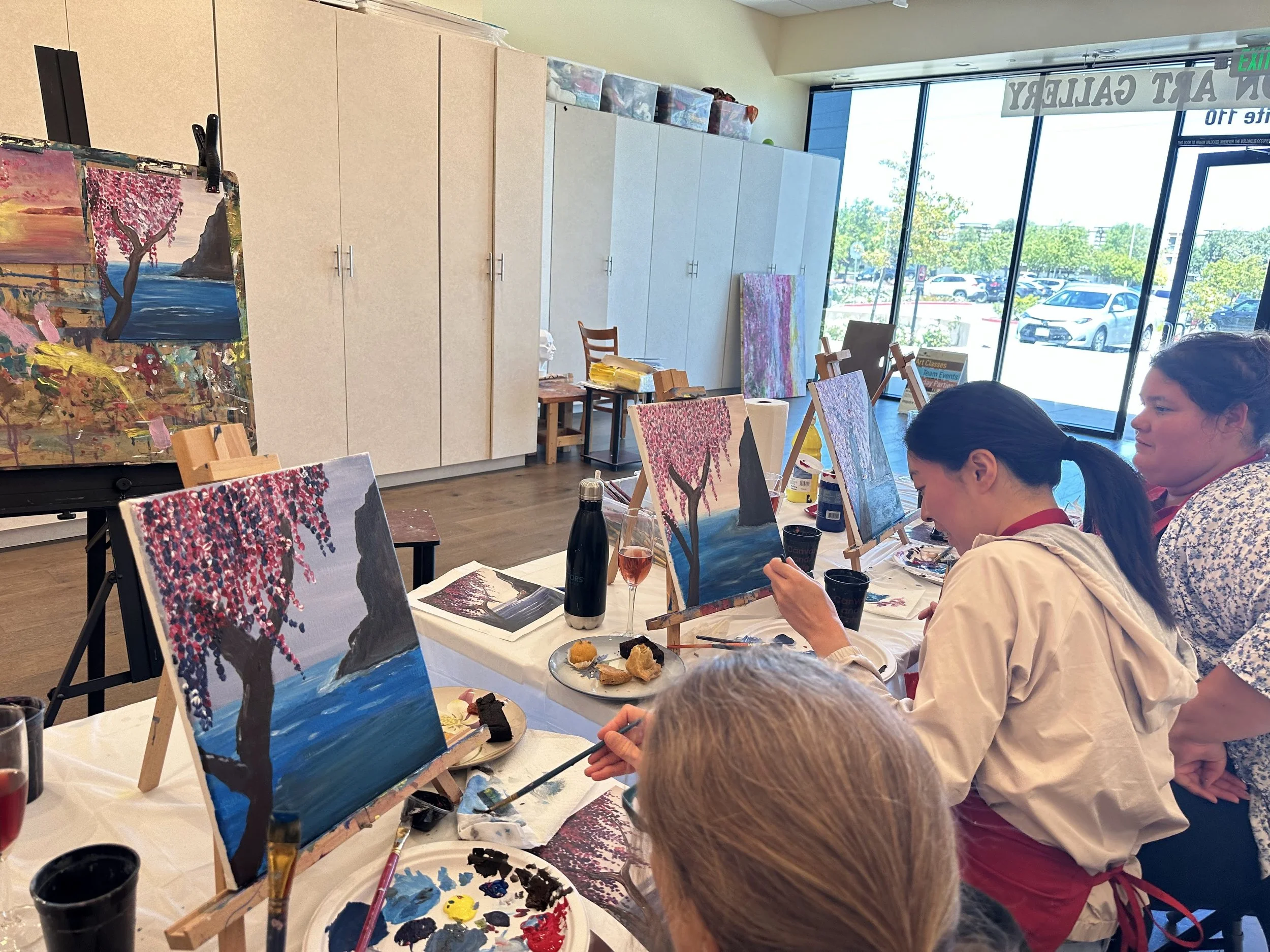 People painting cherry blossom trees on canvases at a paint and sip class in a bright, spacious art studio with large windows and parked cars outside.