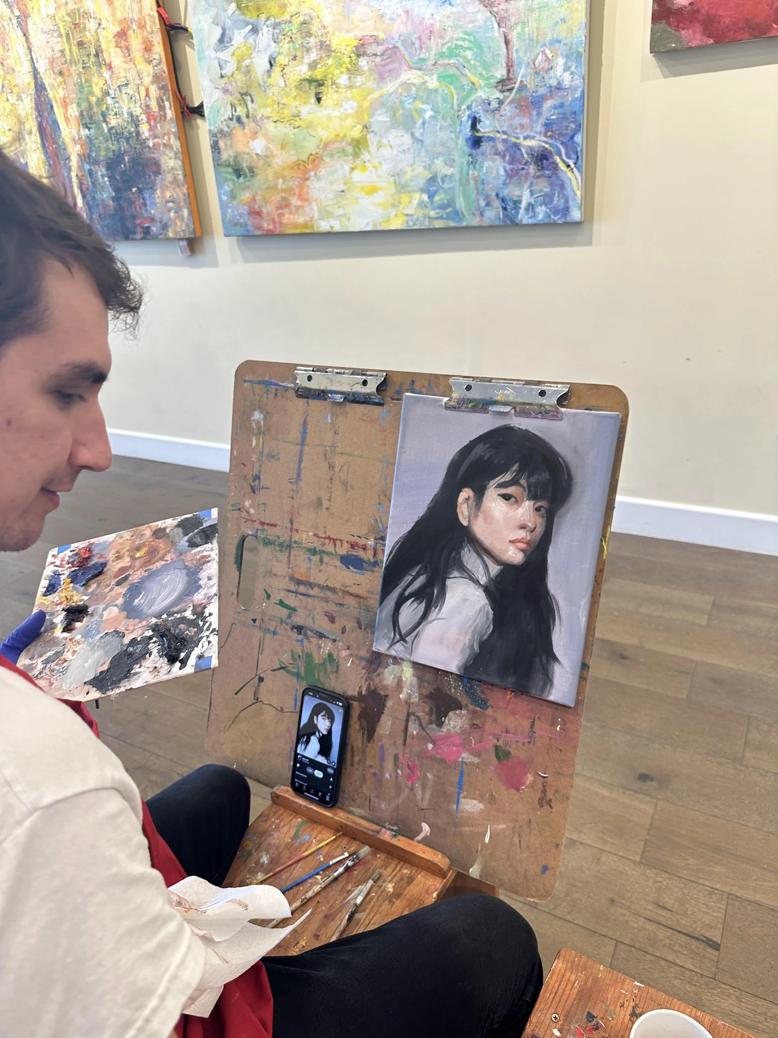 An artist painting a realistic portrait of a woman with dark hair, on canvas, in an art gallery. The artist is sitting on a stool, holding a palette, with a phone displaying the portrait on the stand.