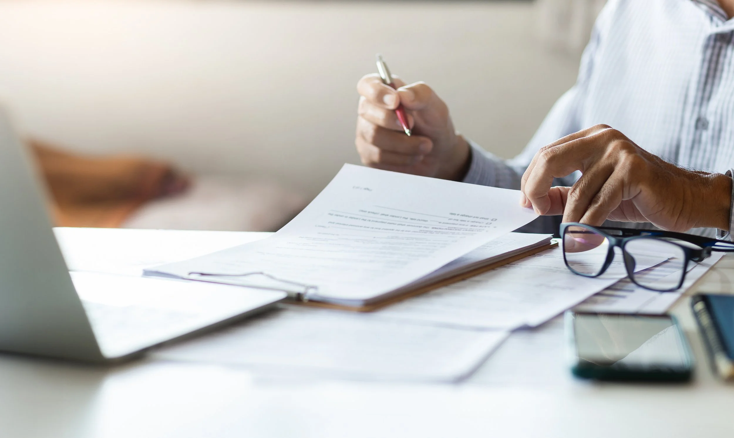 Close-up of a person in a striped shirt reviewing a document on a table with glasses and a notebook, indoors with natural light.