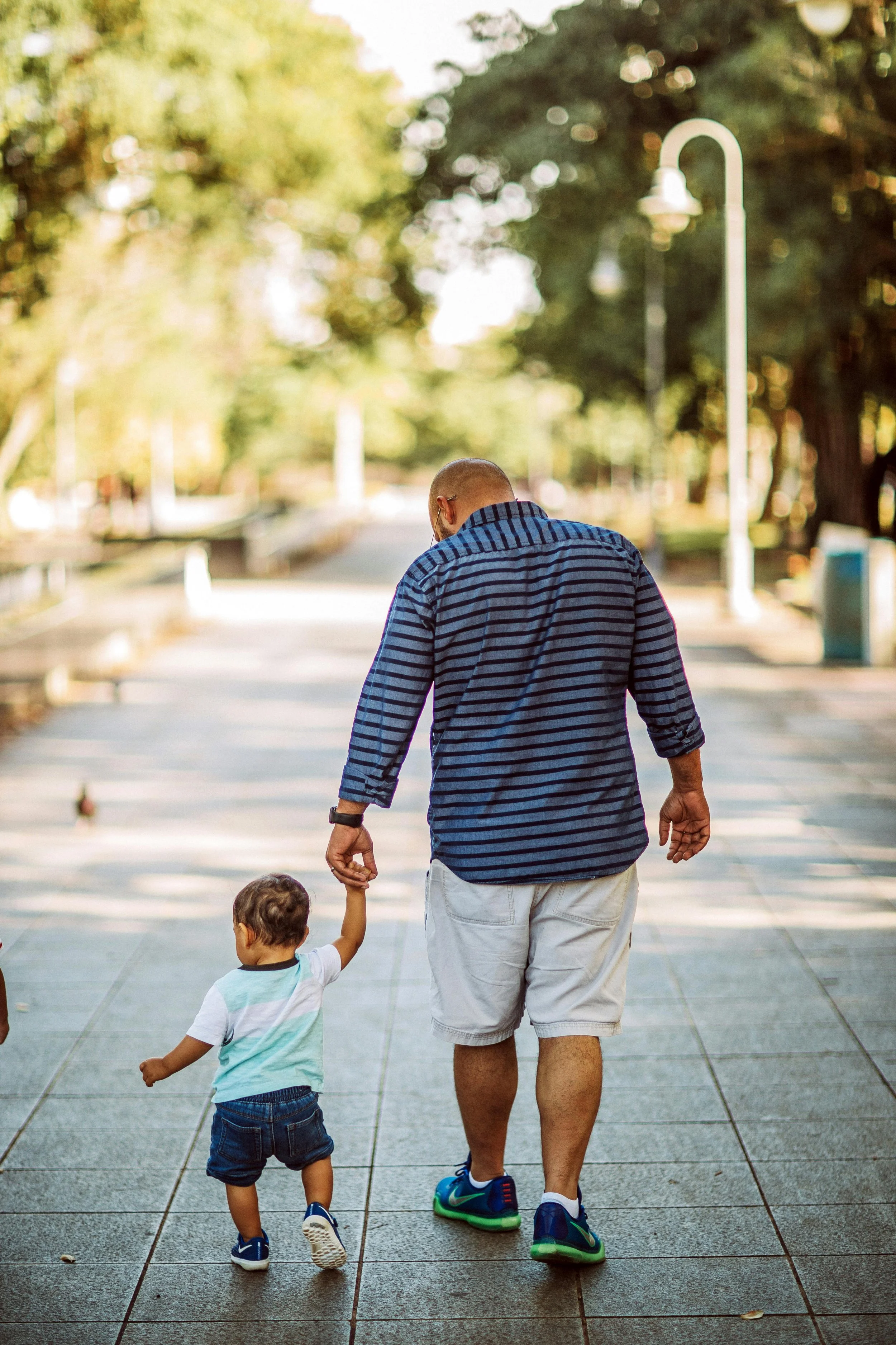 Man and young boy walking hand in hand outdoors on a sunny day, walking on a paved pathway lined with trees and streetlights.