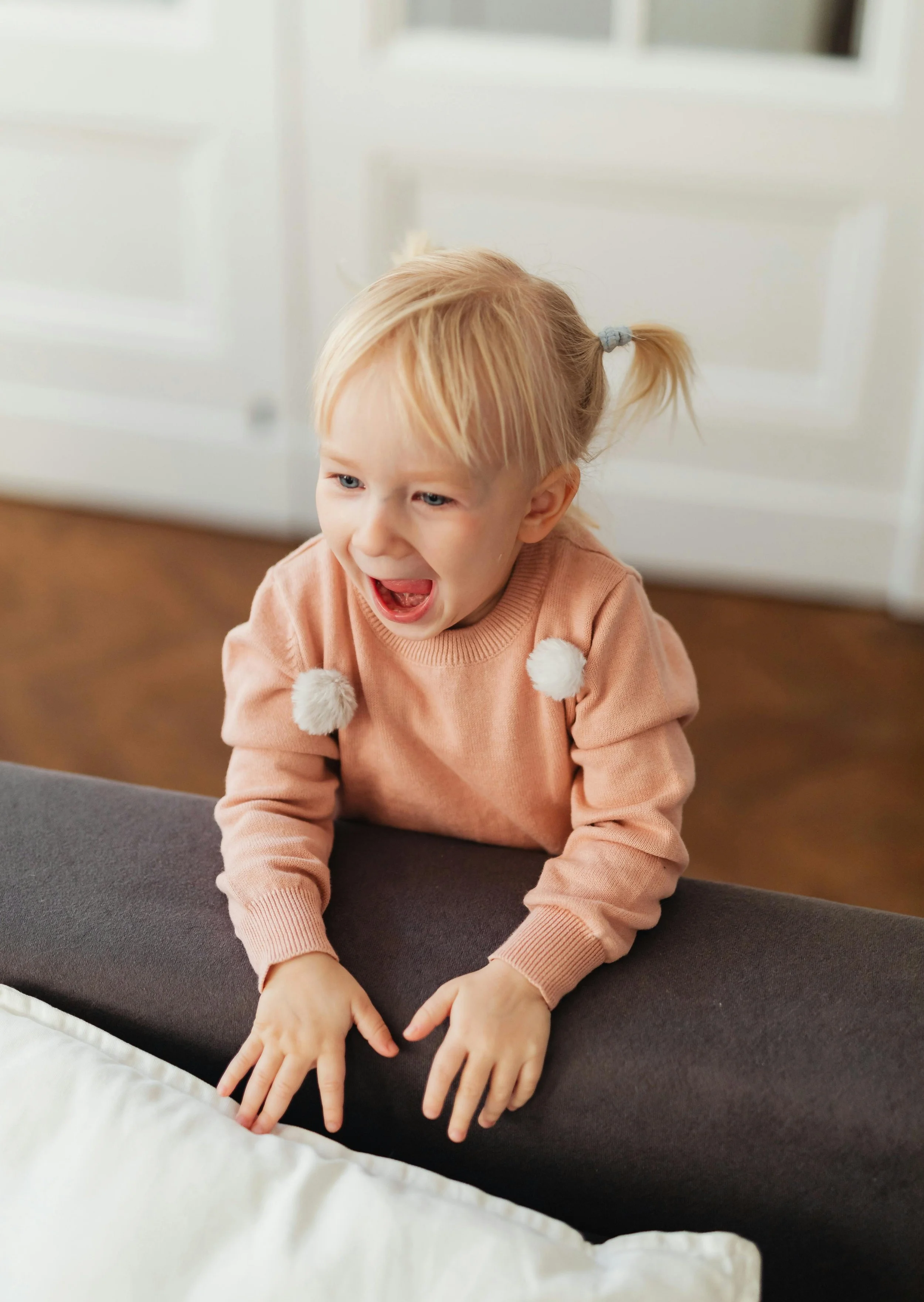 A young Autistic girl with blonde hair in a ponytail, wearing a peach-colored sweater with pom-poms, practicing communication at home with a parent out of view, with her hands and mouth open, looking down.