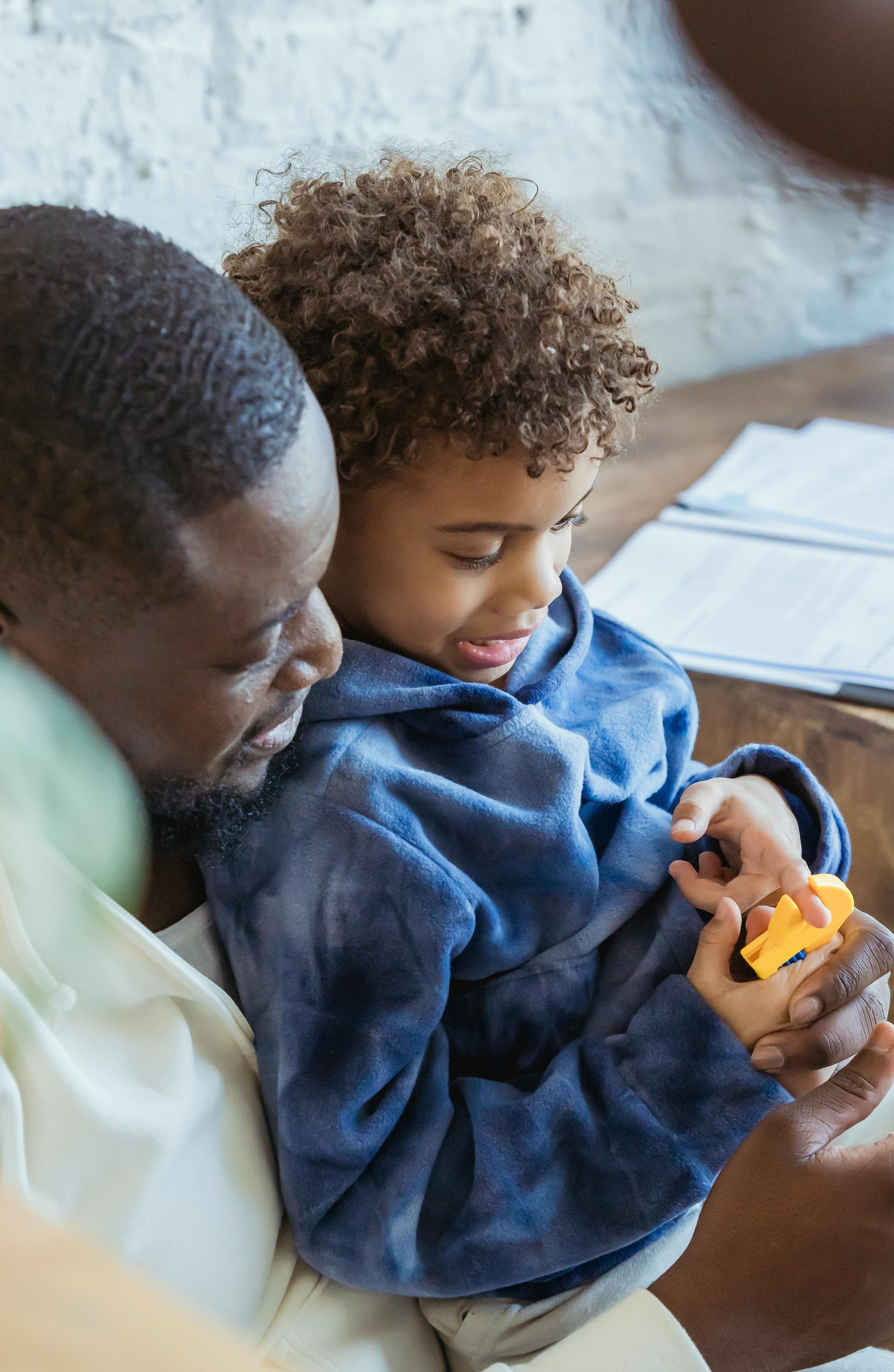 A man and a boy sitting close together, looking at a toy the boy is holding. The boy has curly hair and is wearing a blue hoodie. The man is wearing a light-colored shirt and is assisting or sharing in the moment.