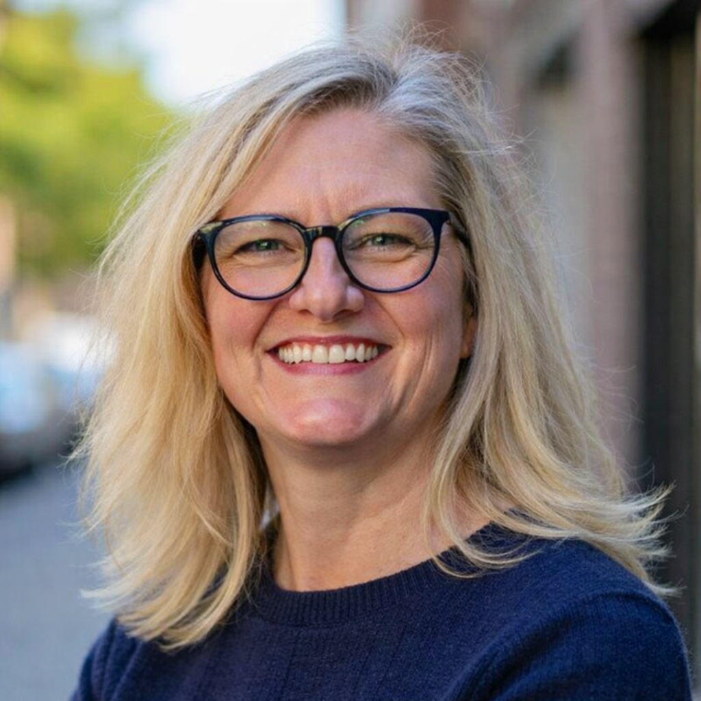 Close-up of a woman with blonde hair and glasses smiling outdoors.