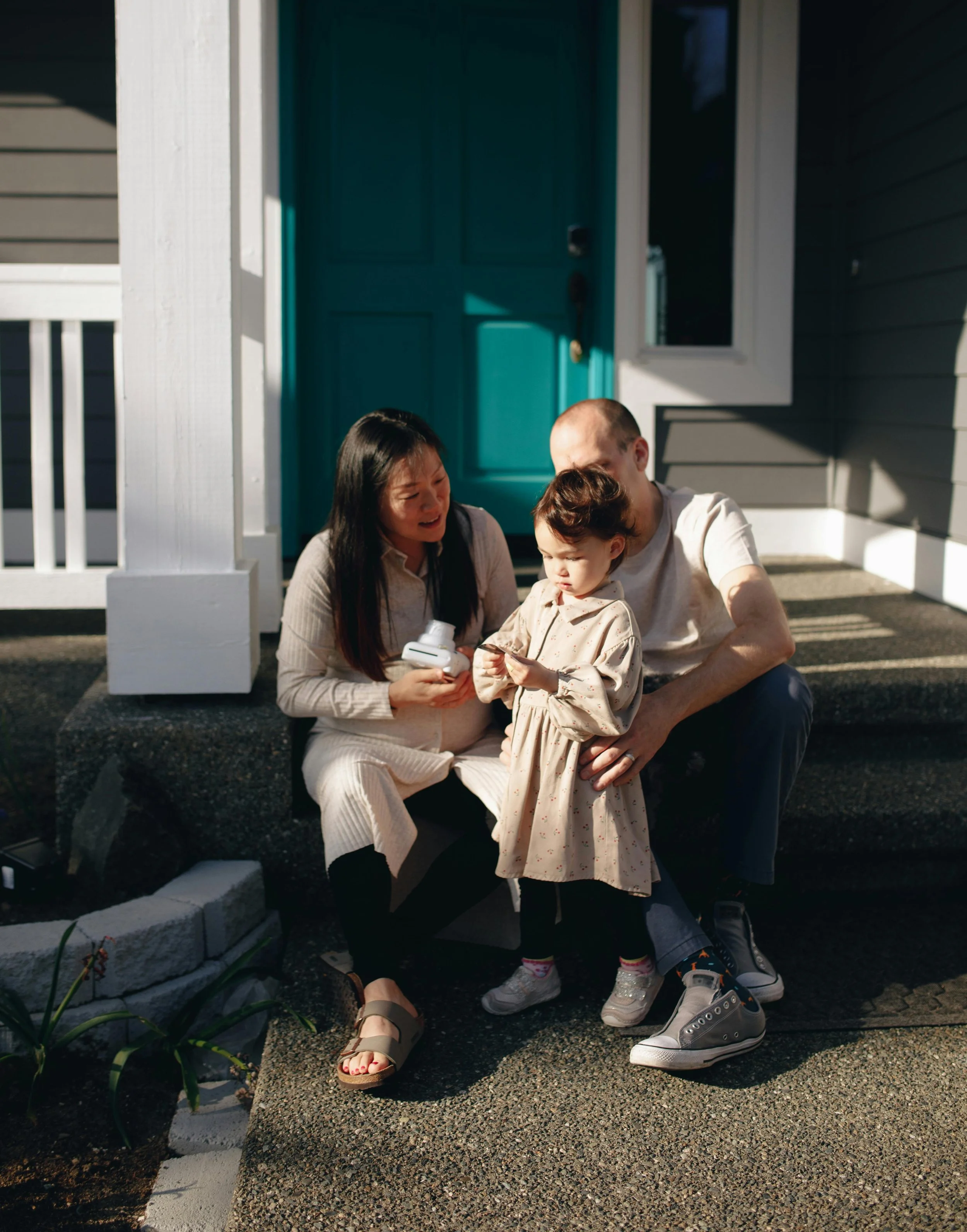 A family of three calmly sitting on steps outside a house, looking at a phone together, with a blue door and window behind them.