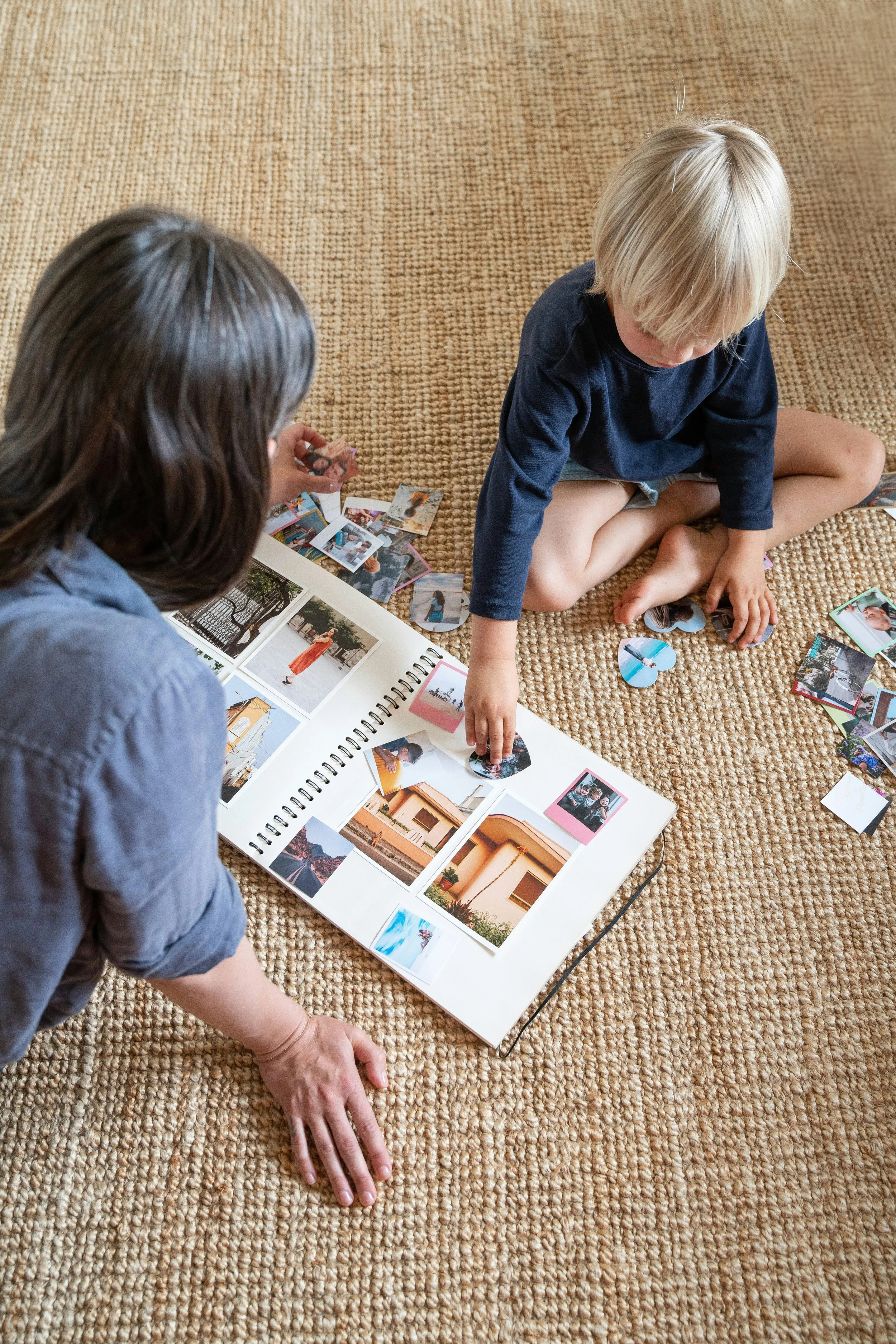 Two young children are sitting on a beige carpet, looking at and organizing photographs in a photo album. One child has dark hair and is wearing a gray shirt, while the other has blond hair and is wearing a navy blue shirt.