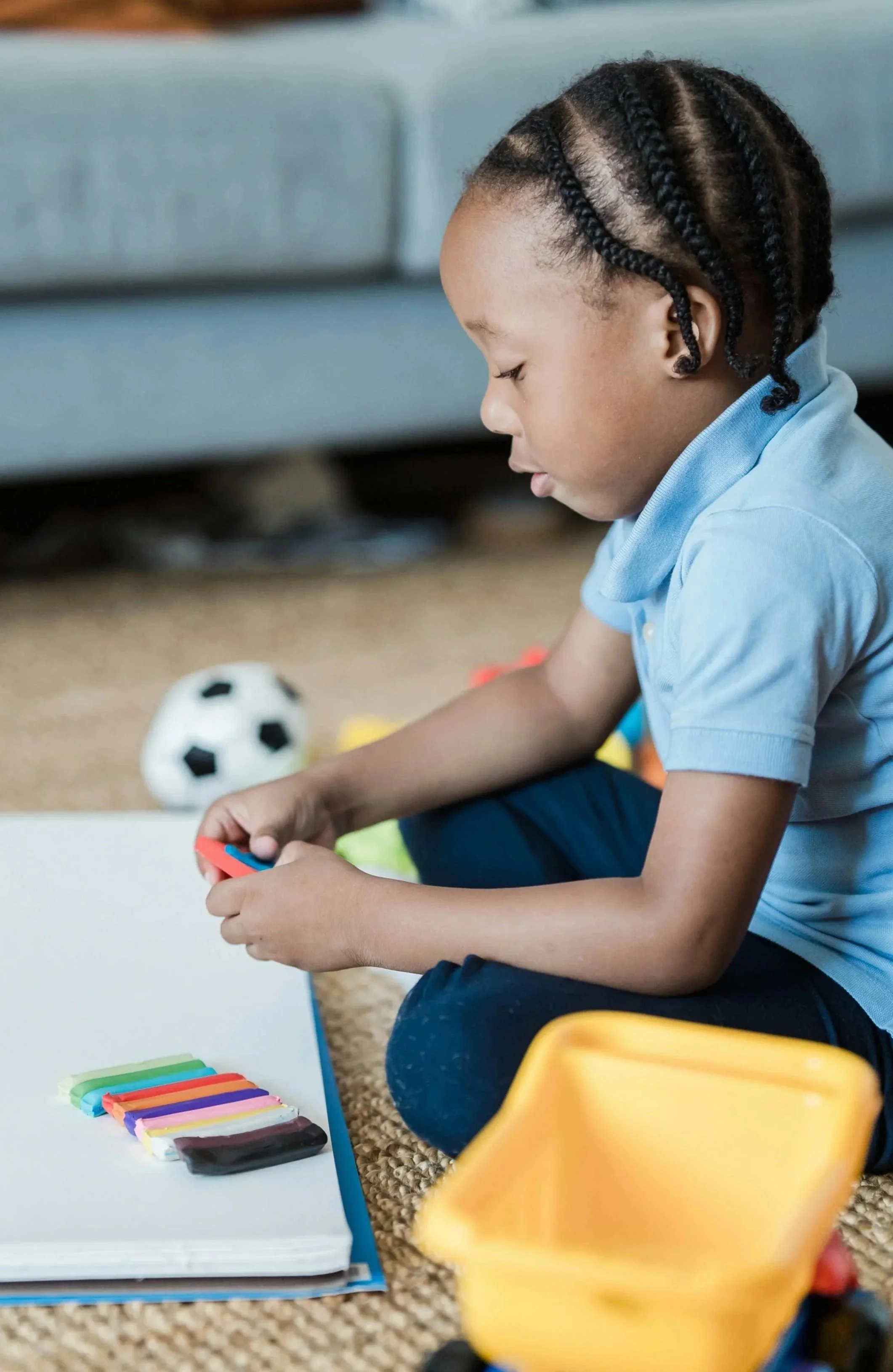 A young boy with braided hair sitting on a carpeted floor, playing with colorful modeling clay, with a soccer ball and a yellow container nearby.