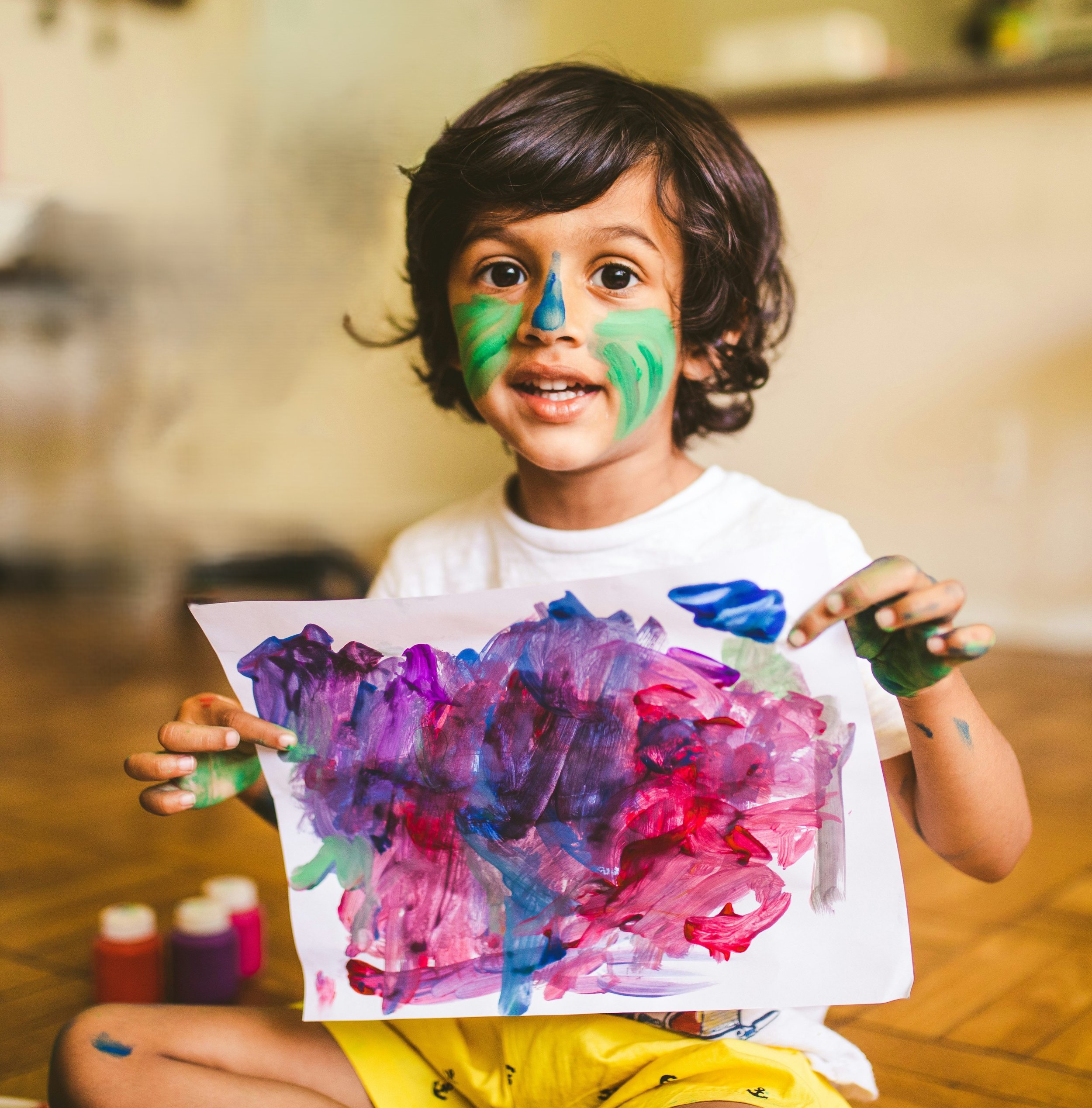 A young child with dark brown hair holds a colorful painting, with face paint resembling a blue nose and green cheeks.