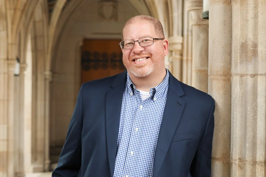 Man in a dark blue suit and glasses smiling while leaning against a stone wall in an arched corridor