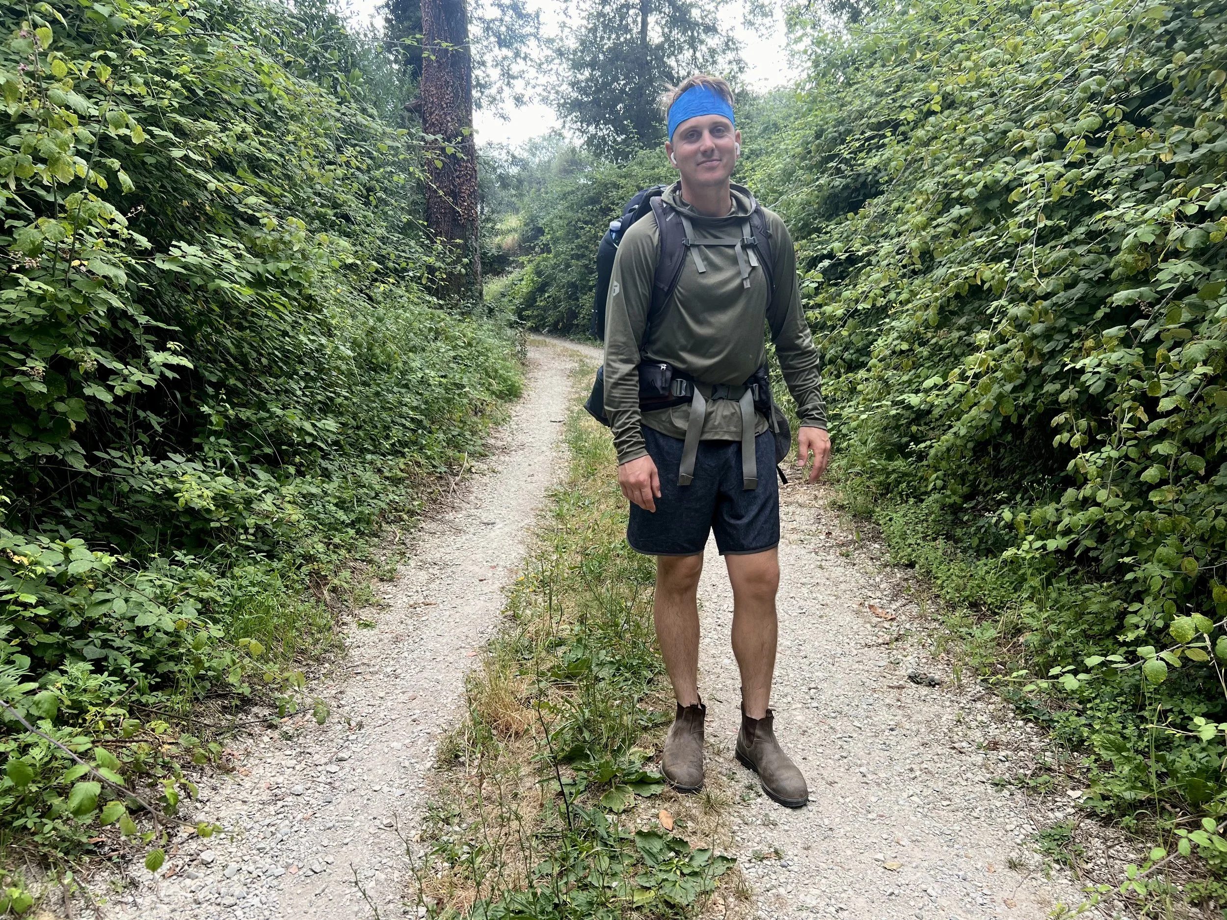 A man standing on a dirt forest trail wearing a blue headband, green long-sleeve shirt, dark shorts, gray hiking boots, and a backpack.