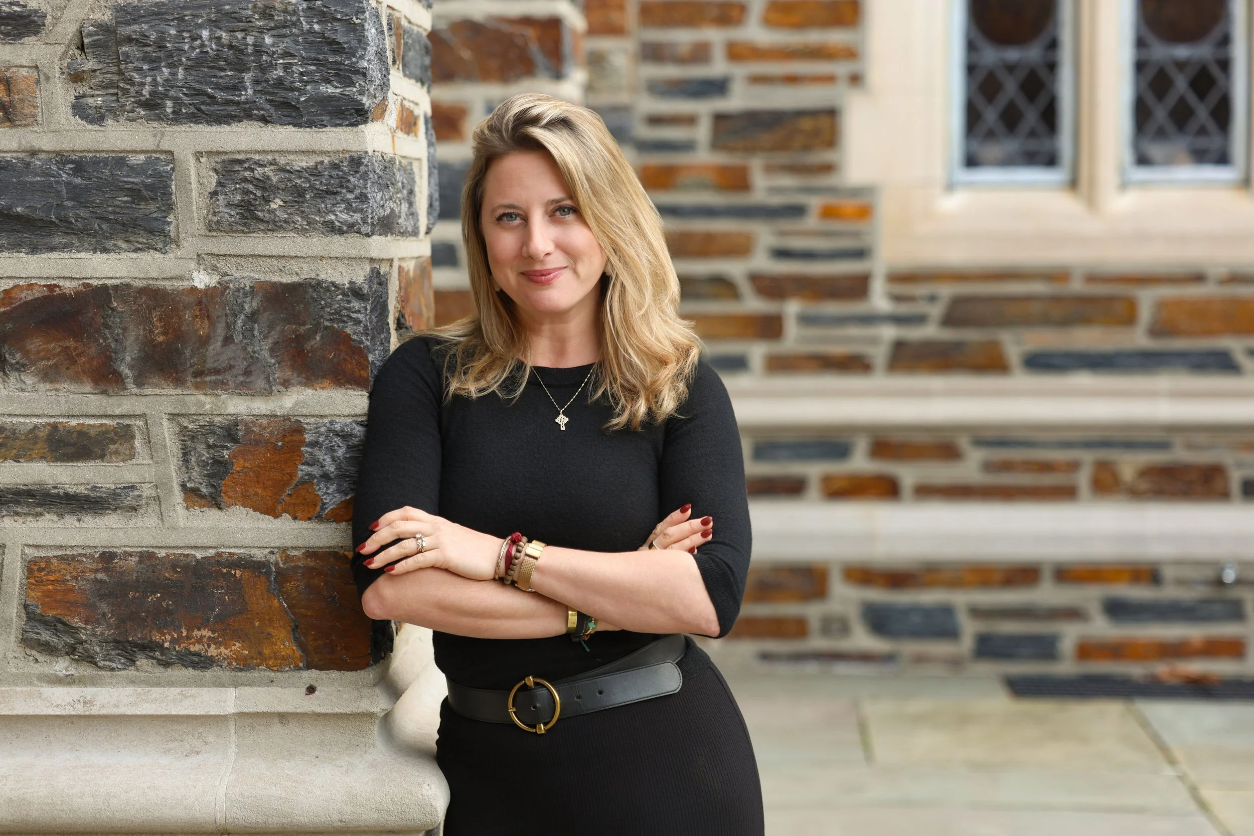 A woman with blonde hair, wearing a black top and belt, stands with her arms crossed in front of a brick and stone wall, smiling at the camera.