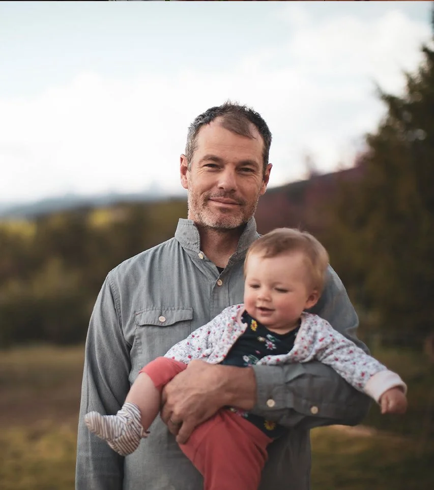 A man holding a young girl outdoors with trees and a cloudy sky in the background.