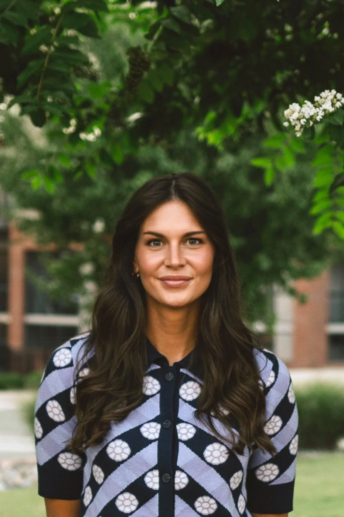 A woman with long brown hair, wearing a black and light purple patterned shirt, standing outdoors with green trees in the background.