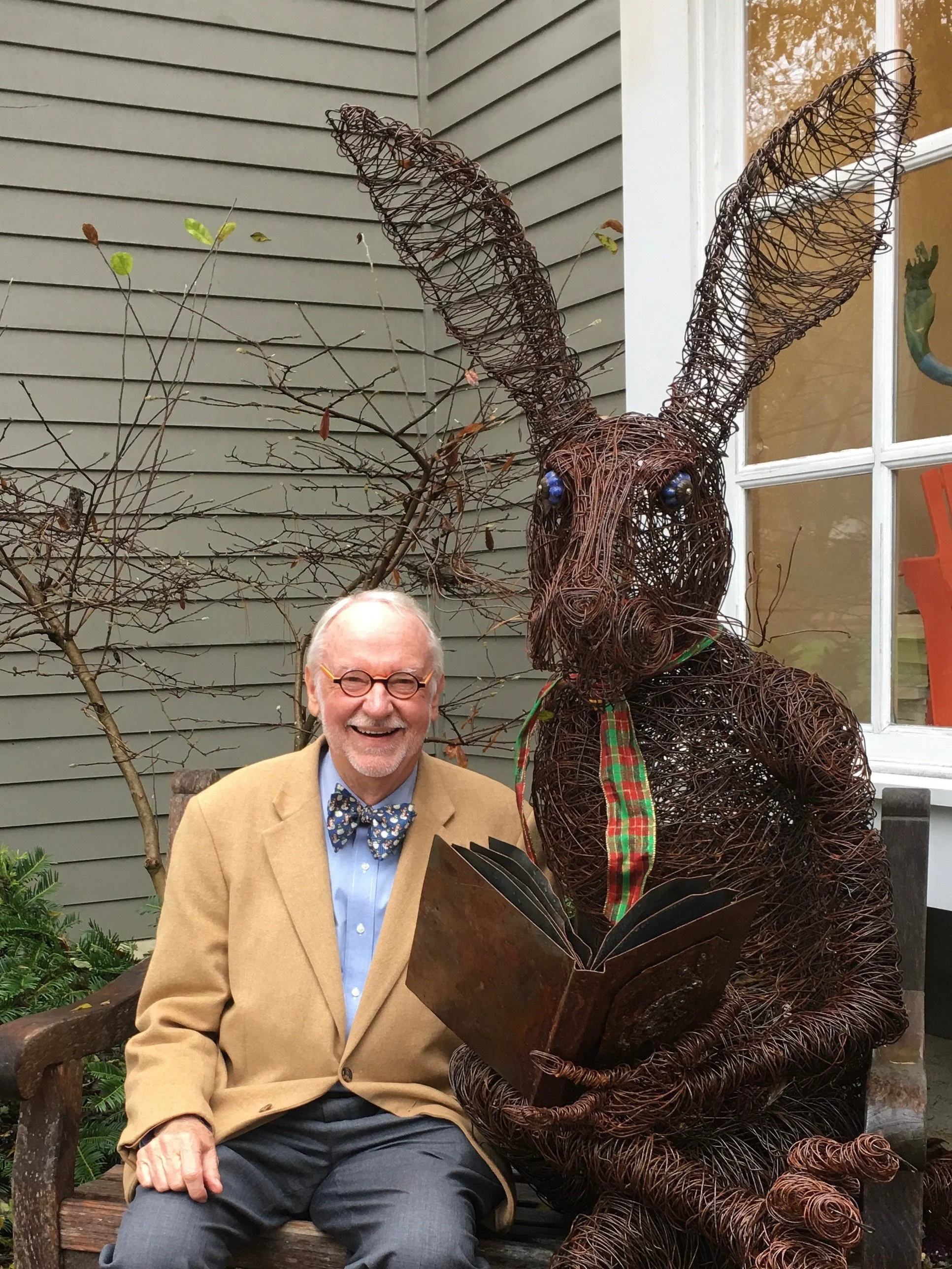 A man with glasses and a bow tie sitting on a wooden bench next to a large bunny sculpture made of wire. The bunny is holding a book and wearing a plaid ribbon or scarf.