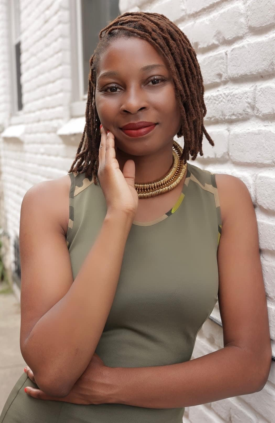 A woman with short dreadlocks wearing a green sleeveless top and a gold necklace, standing against a white brick wall, touching her face gently with her right hand, smiling softly.