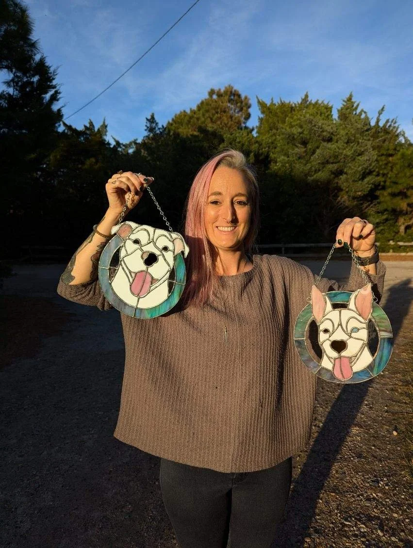 Woman with pink and brown hair holding two stained glass dog face handbags, standing outdoors with trees and a blue sky in the background.