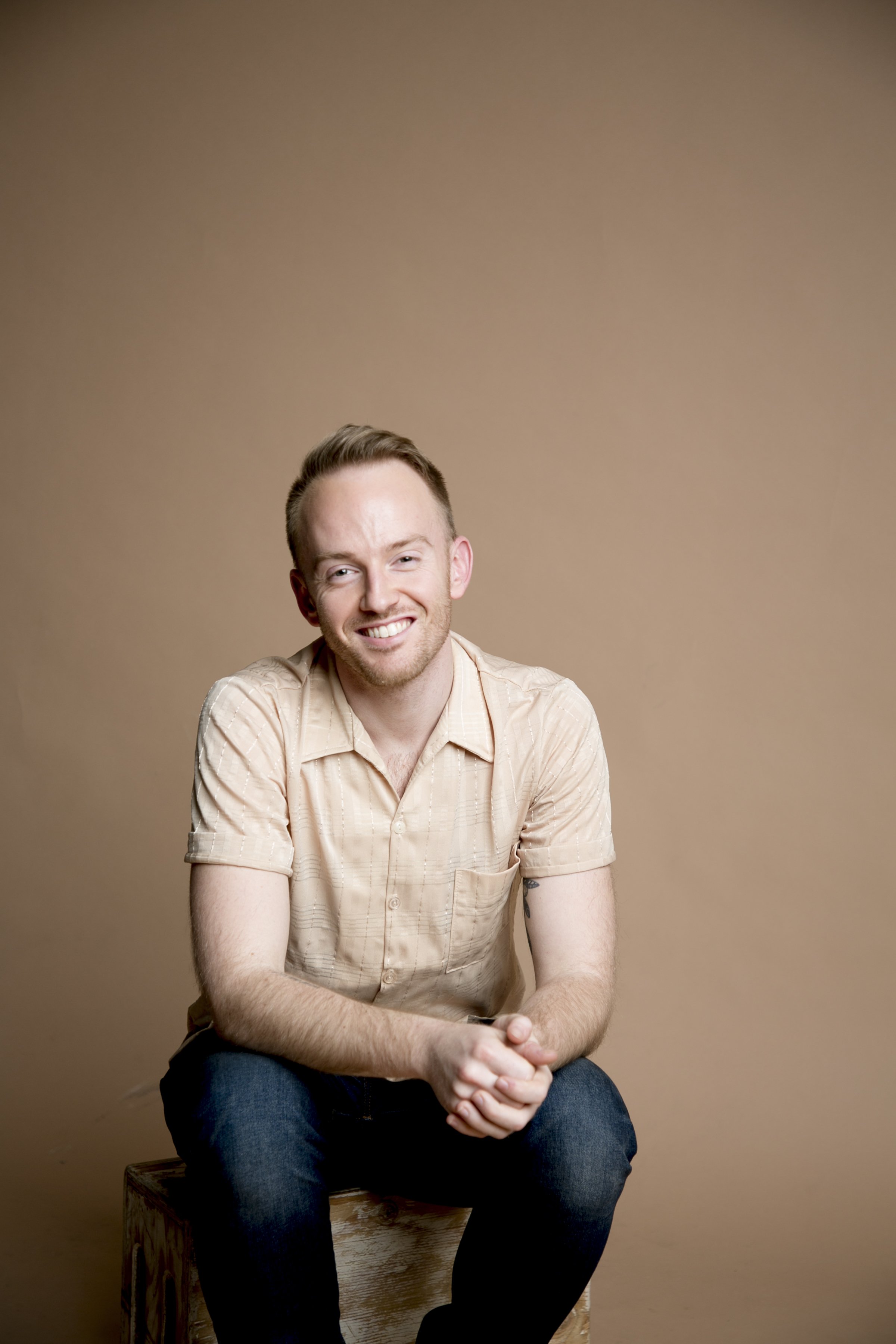 A smiling young man with short, light brown hair and a tattoo visible on his left arm, sitting on a wooden stool against a plain beige background, wearing a short-sleeved, beige plaid shirt and dark jeans.