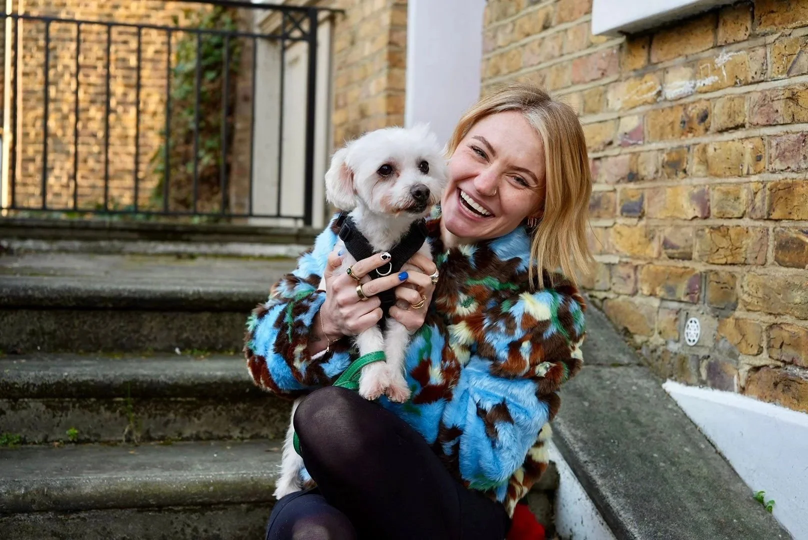A smiling woman with blonde hair wearing a colorful fuzzy jacket crouches on outdoor steps, holding a small white dog with a black harness.