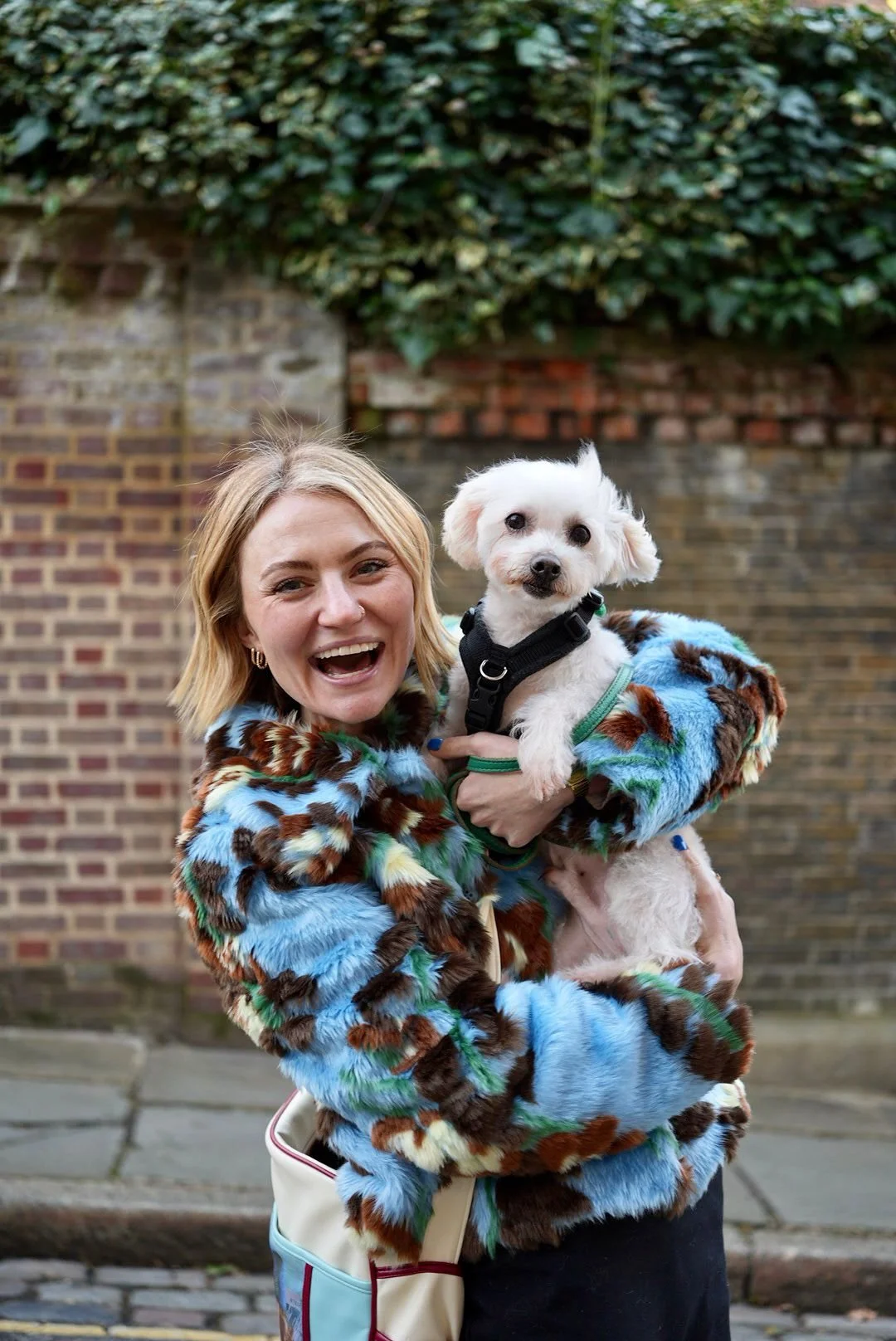 Woman holding a small white dog, both smiling, in front of a brick wall with greenery.