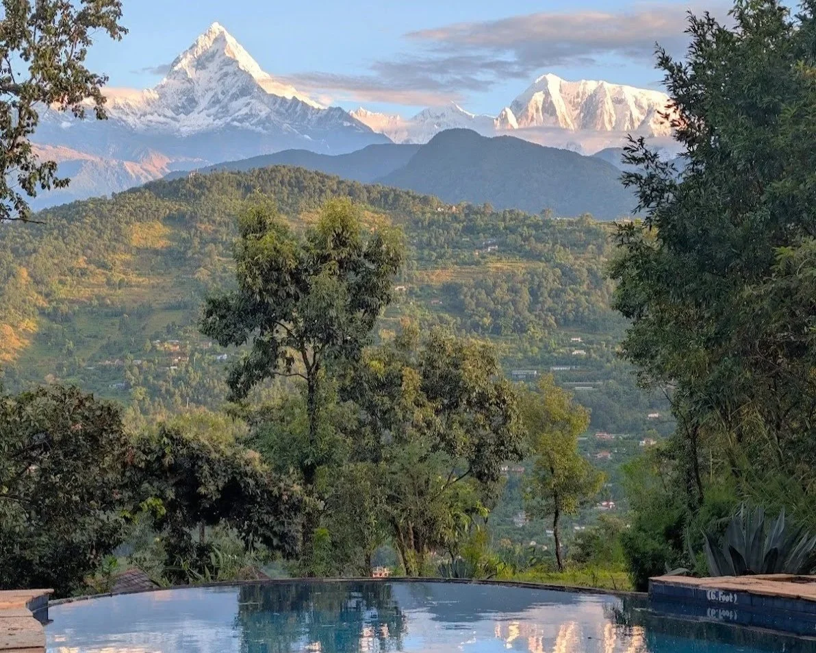 A scenic landscape featuring a swimming pool in the foreground, lush green trees and hills, and snow-capped mountains in the background under a partly cloudy sky.