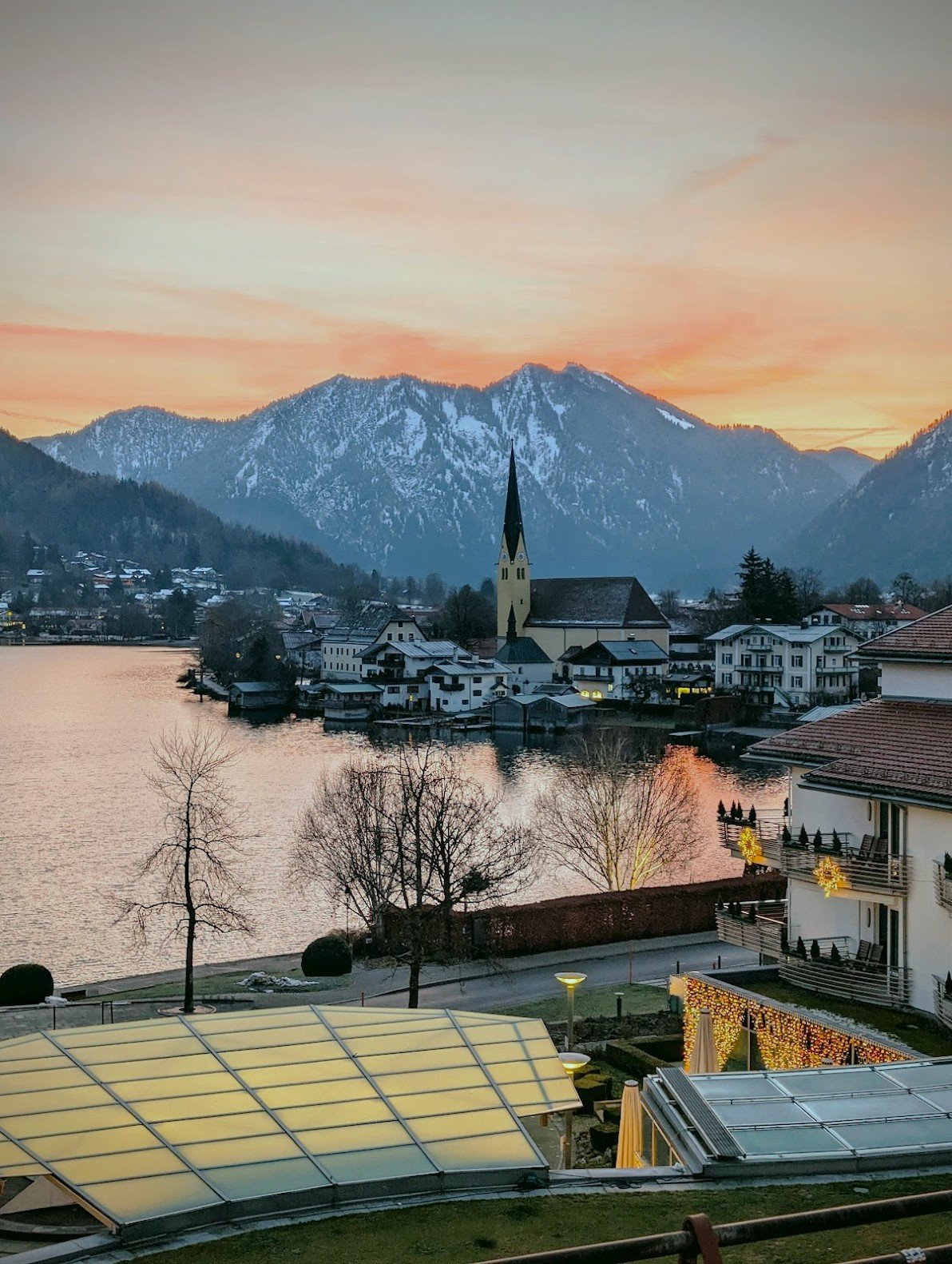 Sunset over a small lakeside town with snow-capped mountains in the background, a church with a tall steeple, residential buildings, and trees.