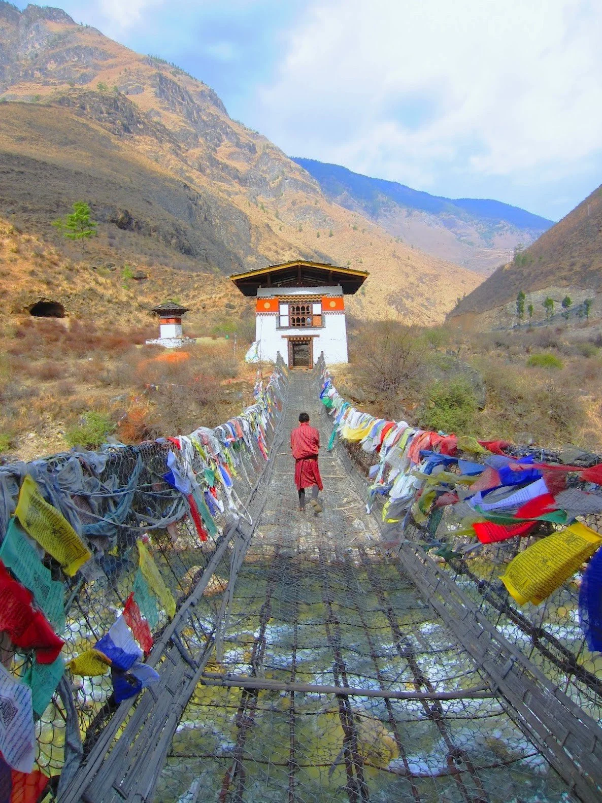 A person walking across a suspension bridge decorated with colorful prayer flags, leading to a traditional structure in a mountainous landscape.