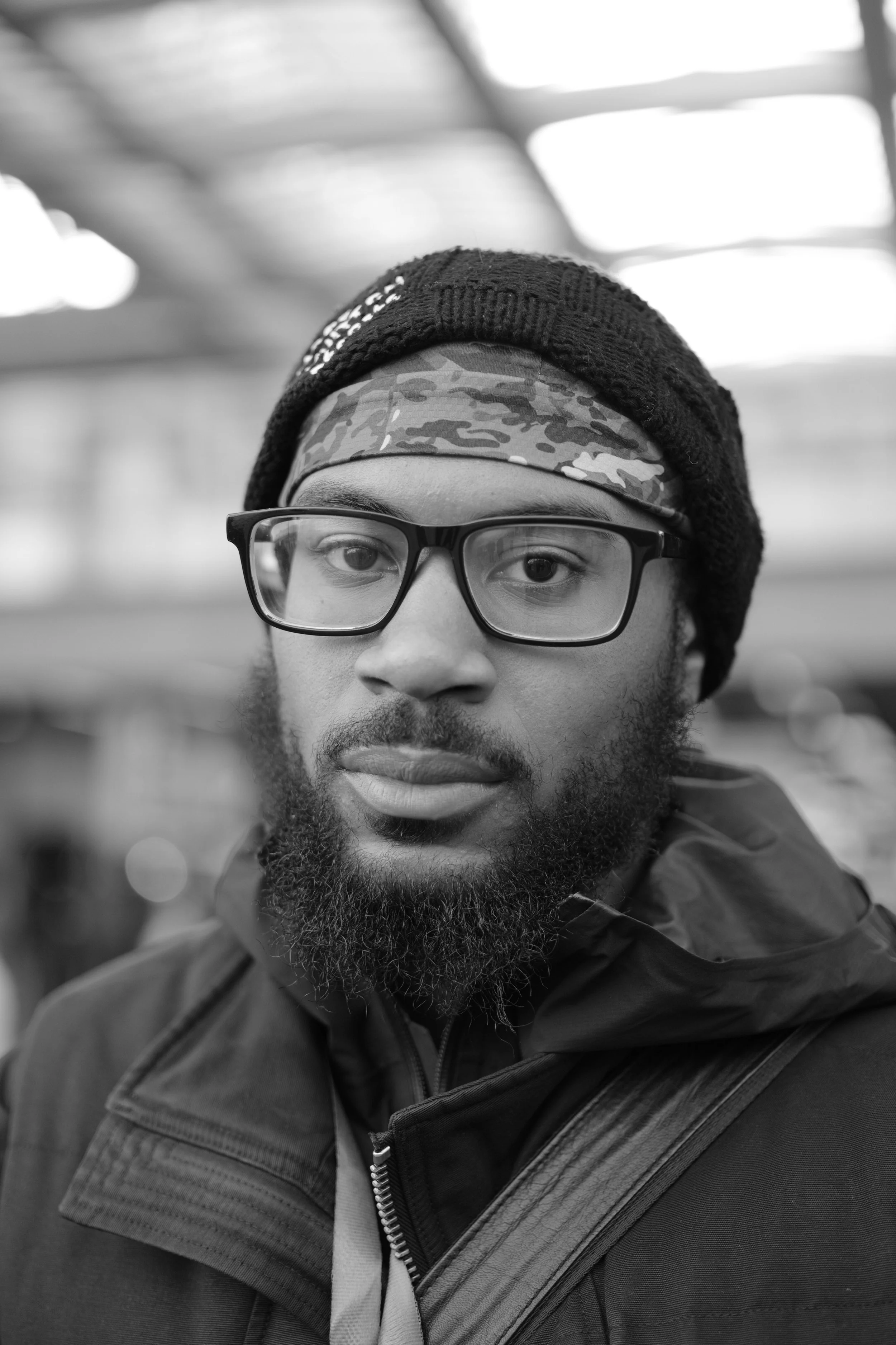 A black-and-white photo of a man with glasses, a beard, wearing a beanie, patterned headband, jacket, and backpack, looking into the camera.