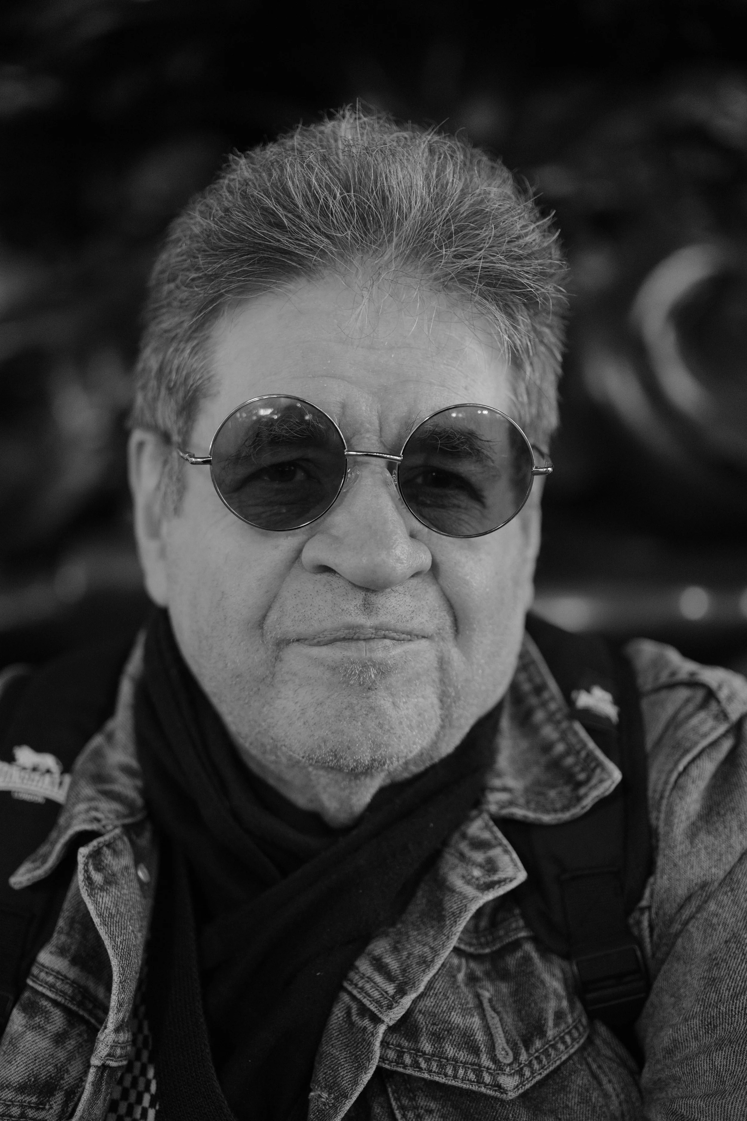Close-up black and white photo of a man wearing round sunglasses, denim jacket, and scarf, with a slight smile.