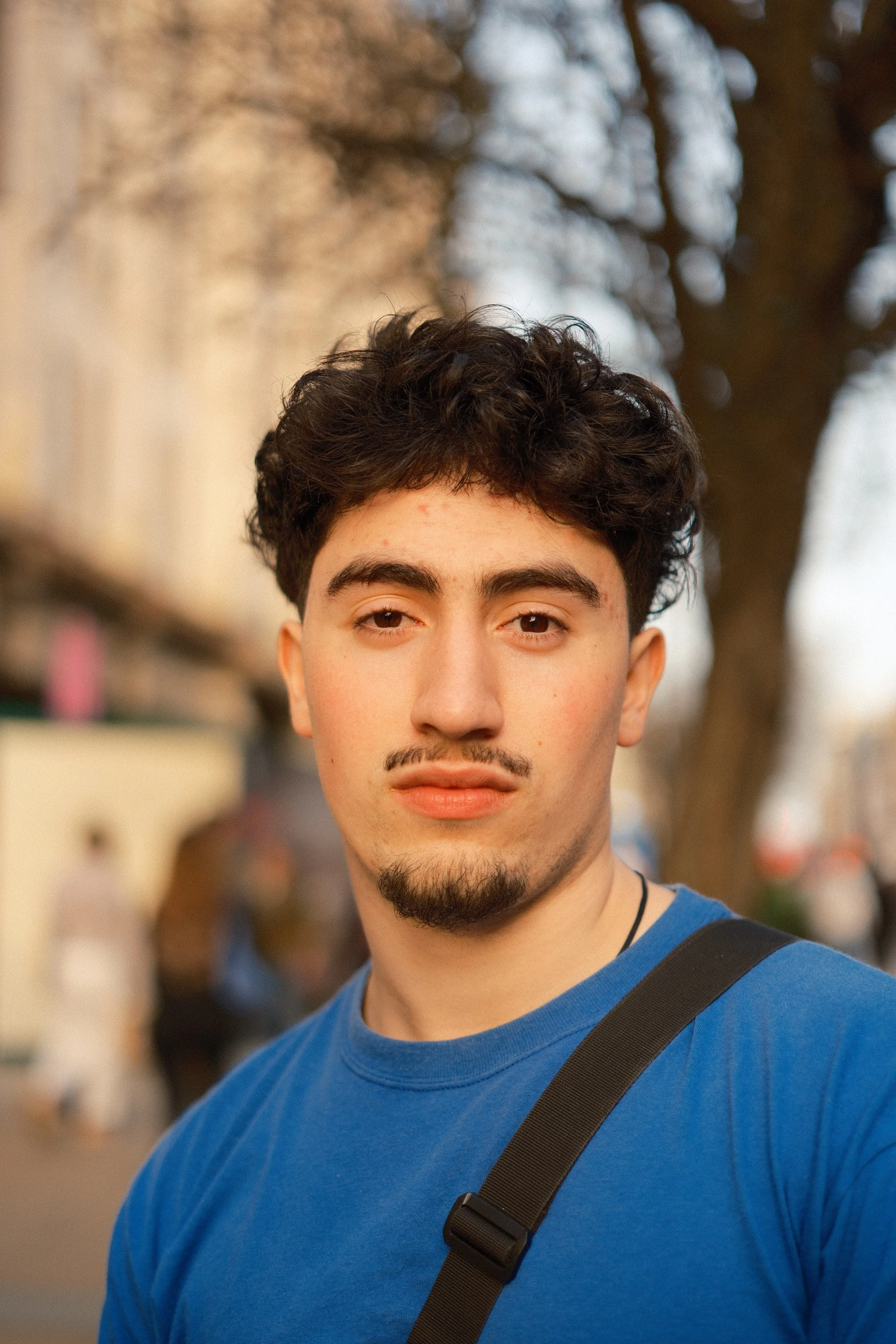 A young man with dark curly hair, a goatee, and light skin, wearing a blue shirt and a black strap, stands outdoors in front of a blurred background including trees and buildings.