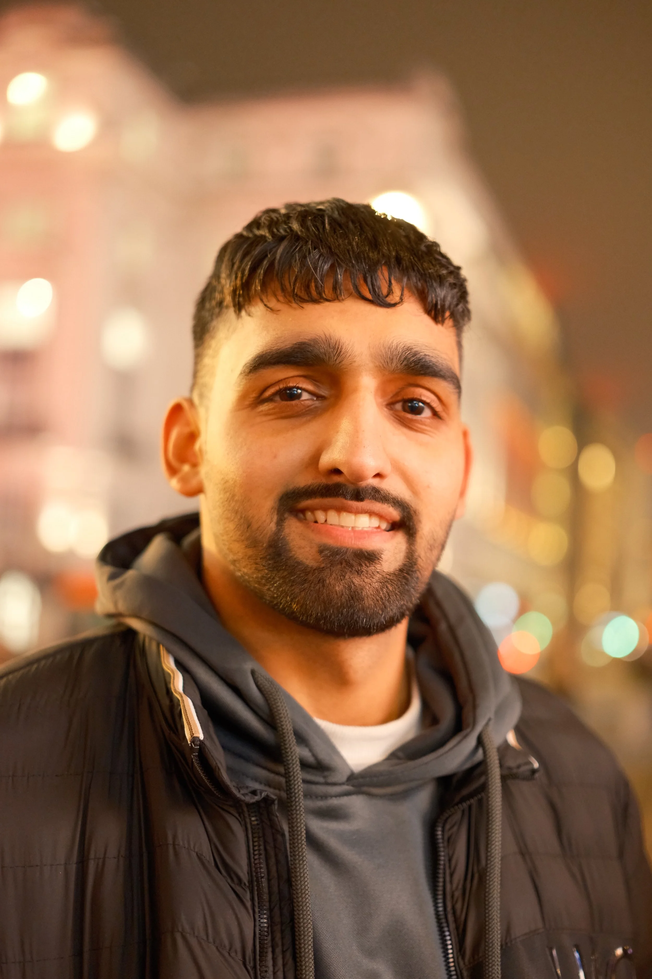 Close-up of a smiling man with dark hair and a beard, wearing a black jacket and gray hoodie, standing outdoors at night with blurred city lights in the background.