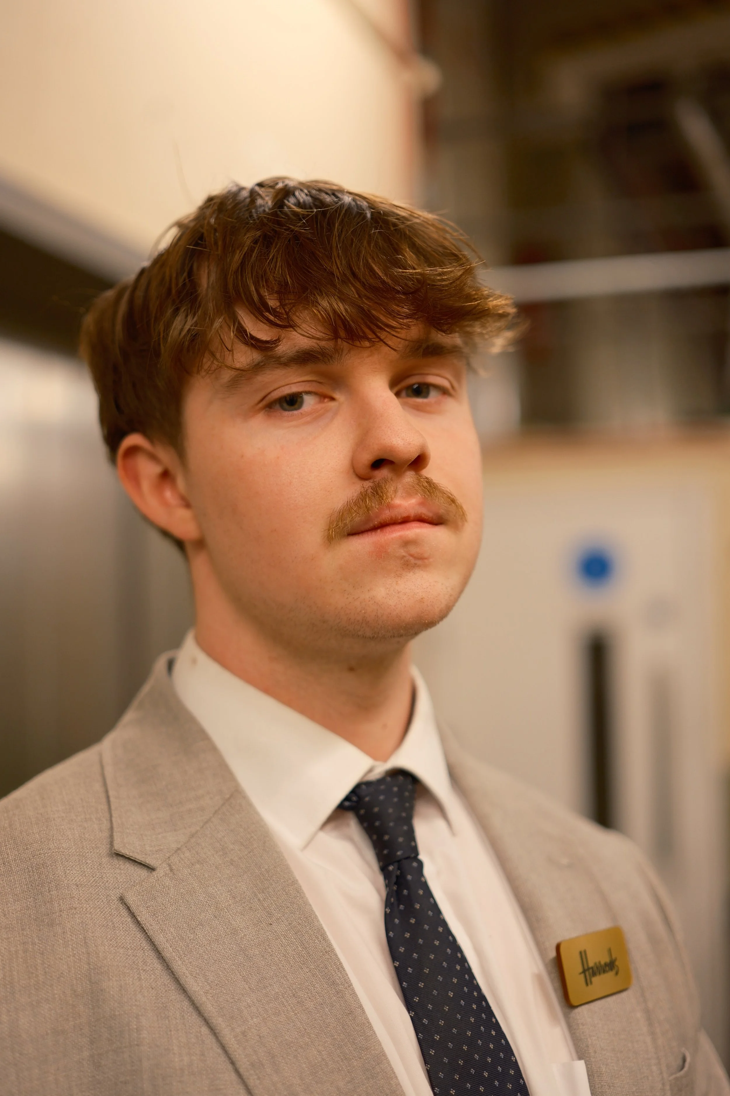 A young man with wavy brown hair, a mustache, wearing a light gray suit, white shirt, dark dotted tie, and a name tag that says 'Hastings'.