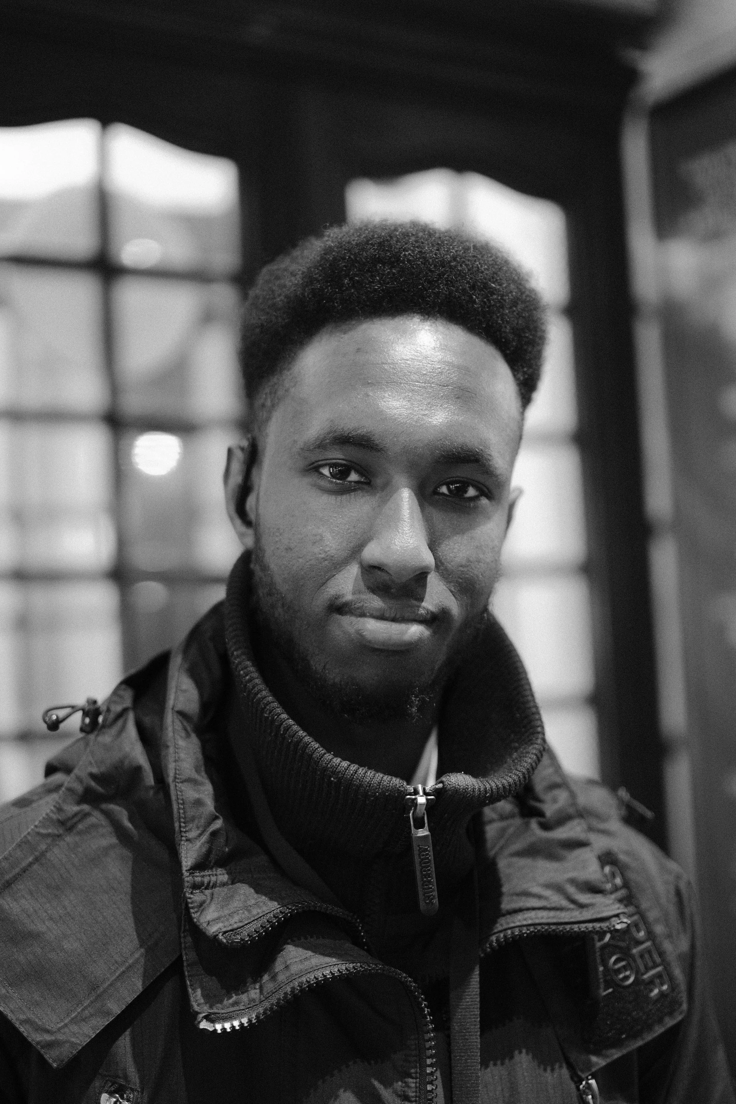 Black and white portrait of a young man with short Afro hairstyle, wearing a jacket with a zippered collar, standing indoors with background windows.