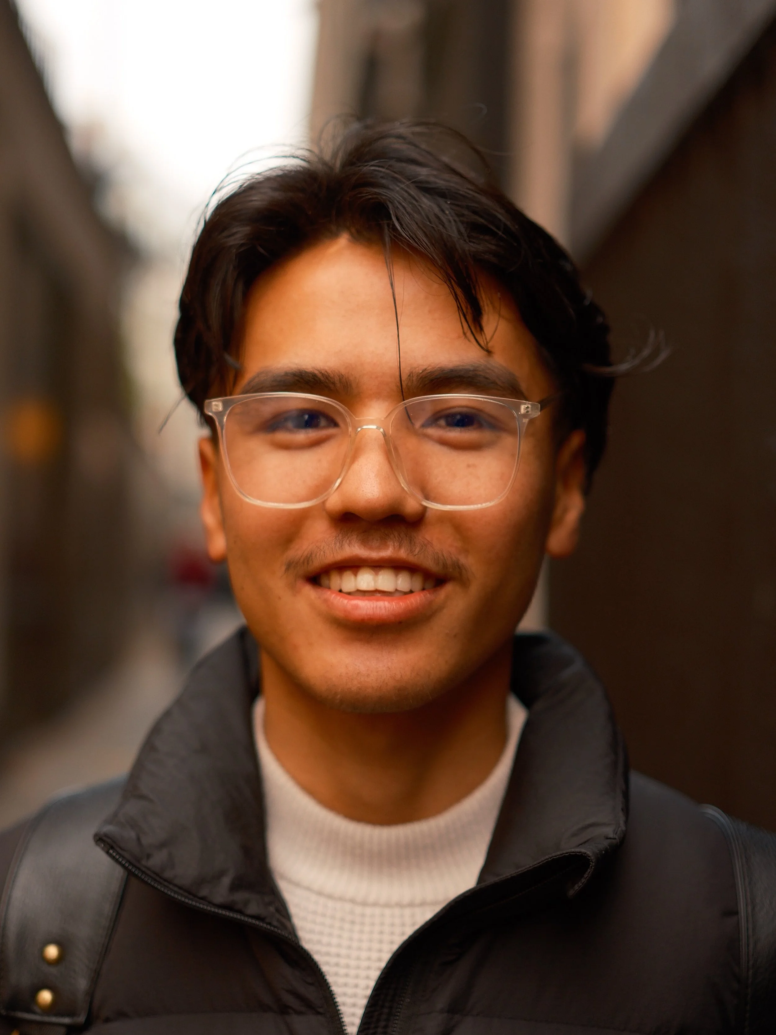 A young man with dark hair, glasses, and a light complexion smiling outdoors in a narrow alleyway.