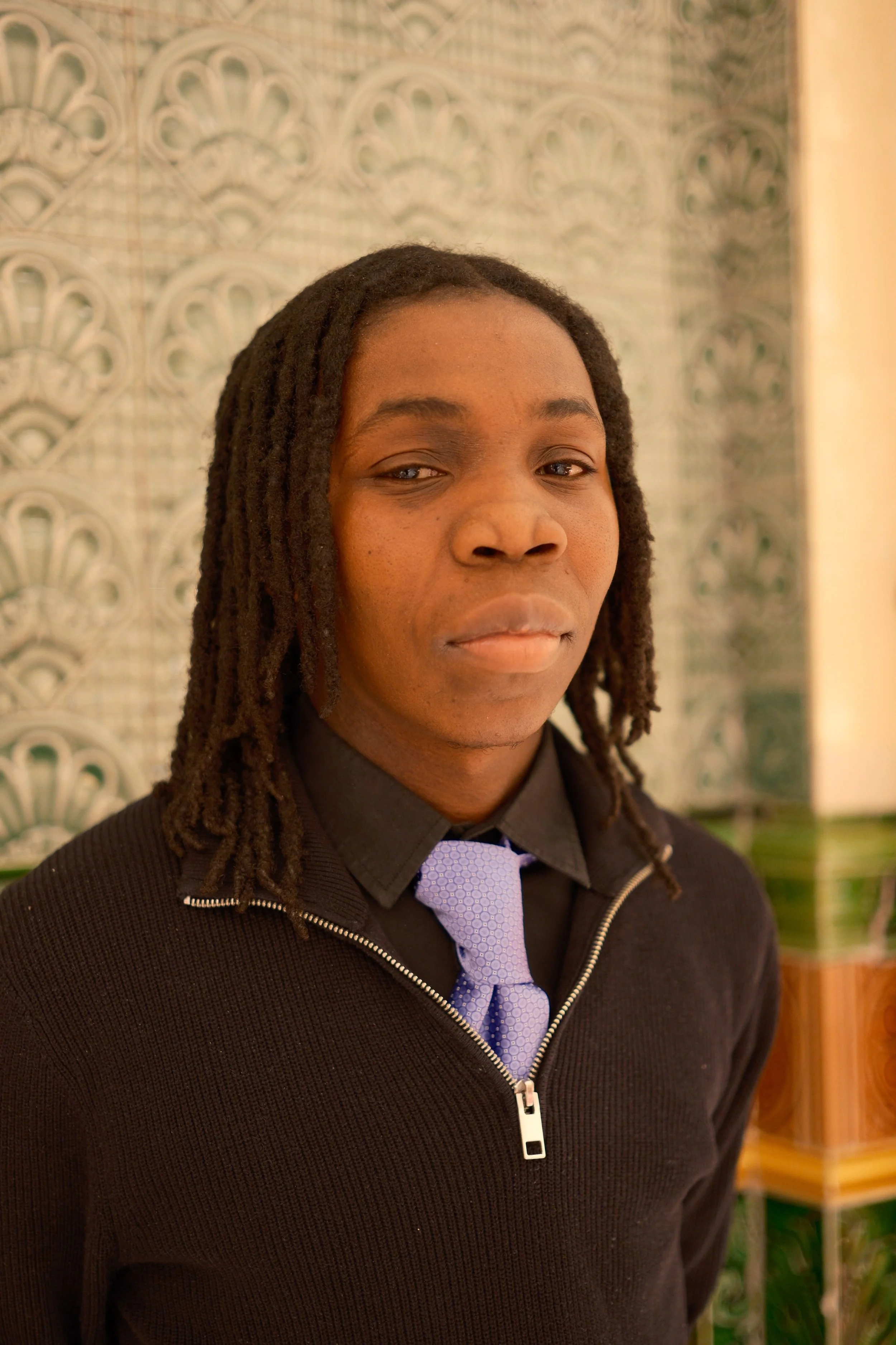 A young man with dreadlocks wearing a black zip-up sweater over a collared shirt and purple tie, standing in front of a patterned wall.