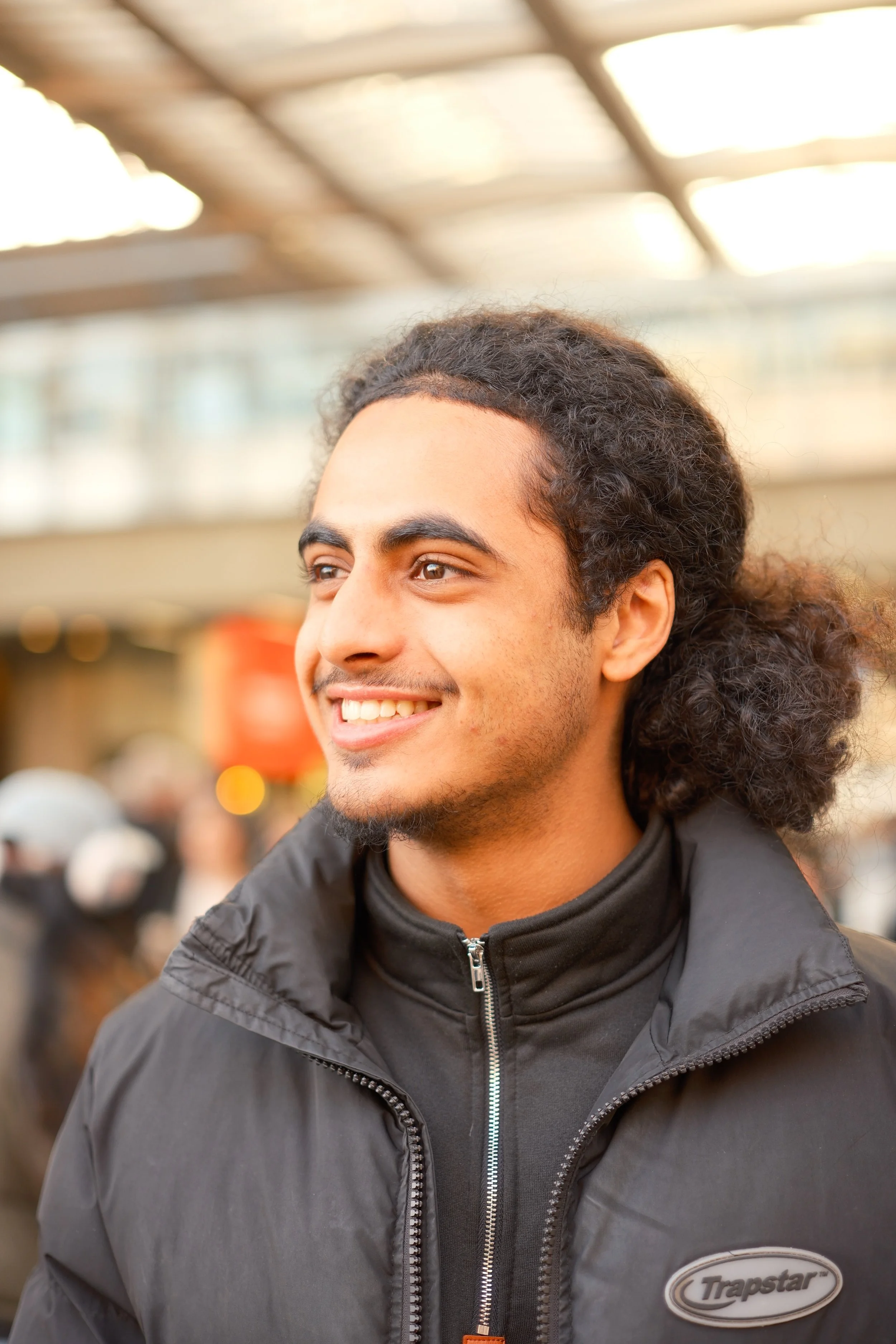 A young man with curly hair smiling, wearing a black jacket, in a busy indoor setting with blurred background.