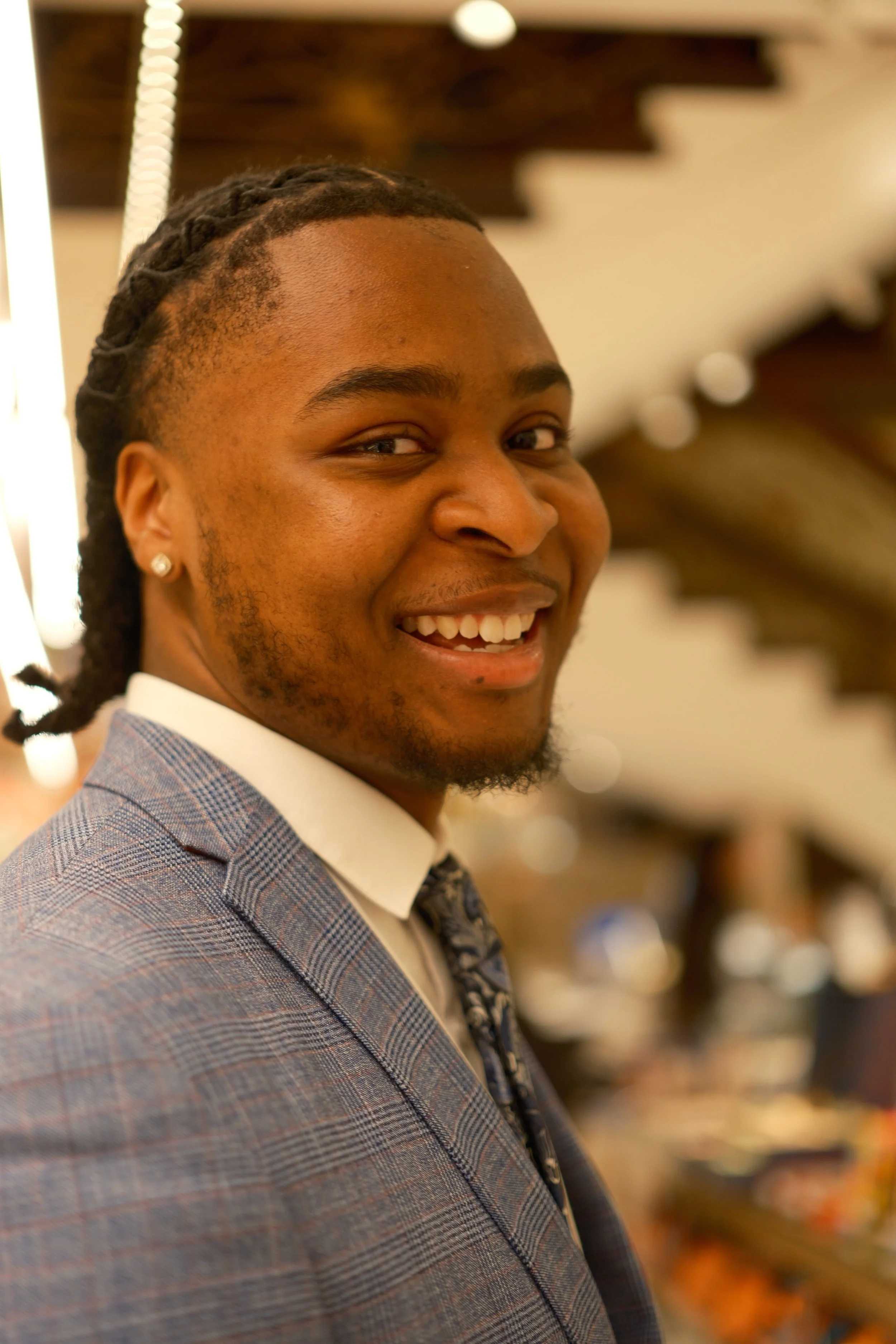 A smiling man with dreadlocks and a beard, wearing a plaid suit jacket, a white shirt, and a patterned tie, in an indoor setting with warm lighting.