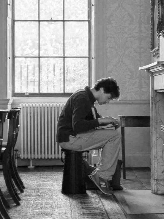 A young man with dark hair sits at a desk near a large window, writing in a notebook.