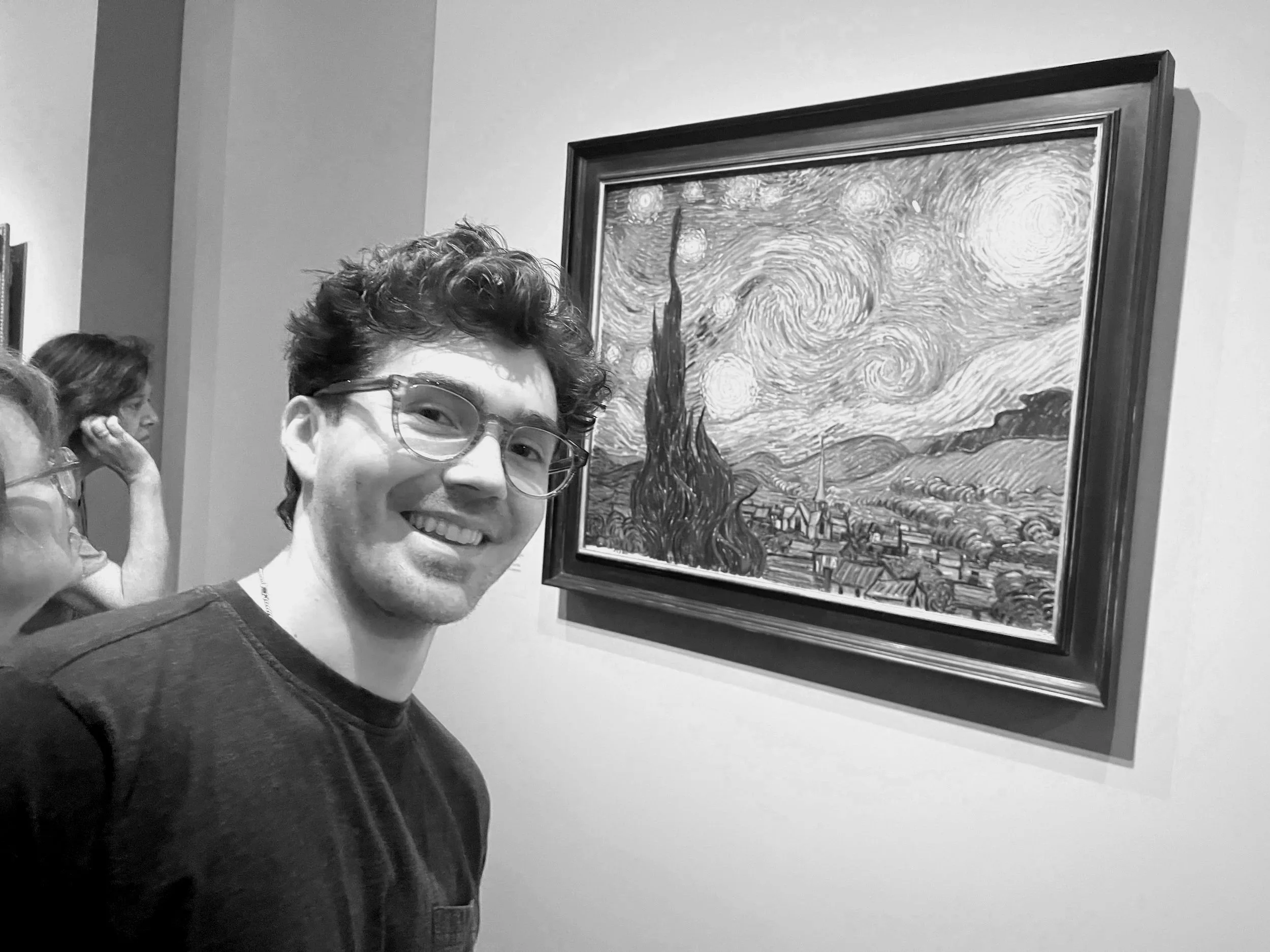 A young man with glasses smiling at an art gallery next to Vincent van Gogh's Painting Starry Night.