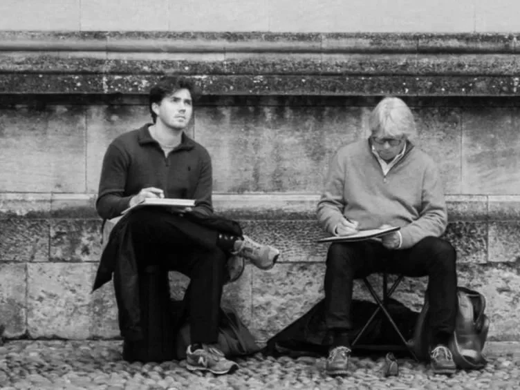 Two people sitting on the sidewalk with notebooks and pens, one looking ahead and the other looking down, against a stone wall.