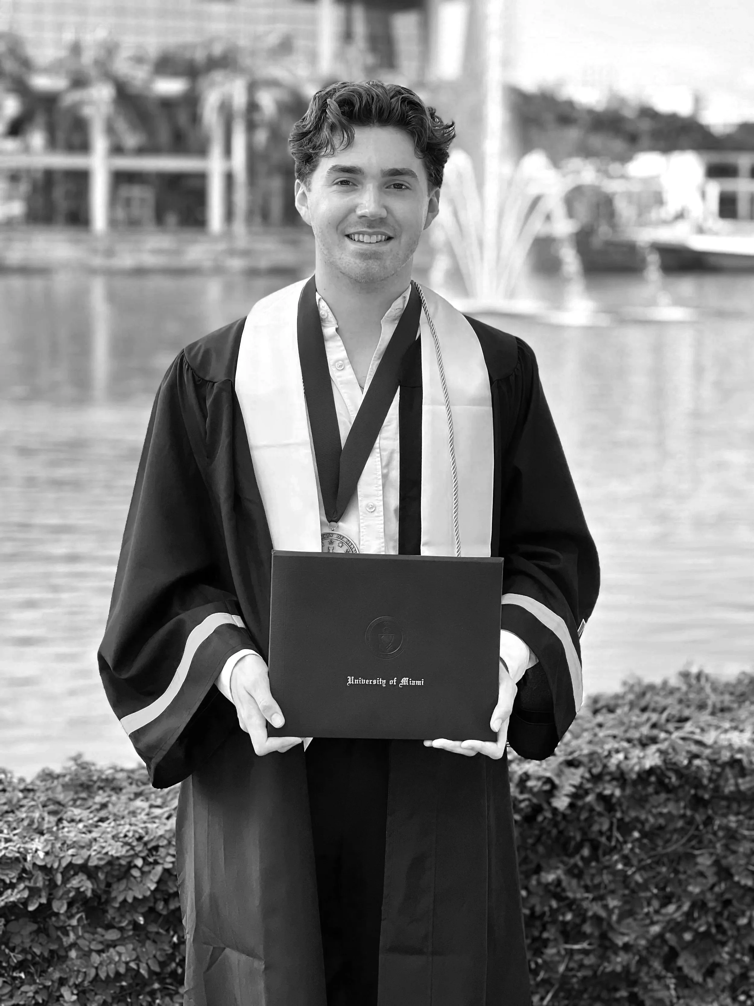 A young man in graduation gown holding a diploma, with a fountain and water in the background.