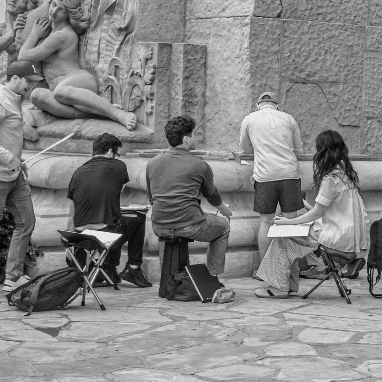 Group of art students sketching and studying a large bas-relief sculpture on a stone wall in a museum or art studio.