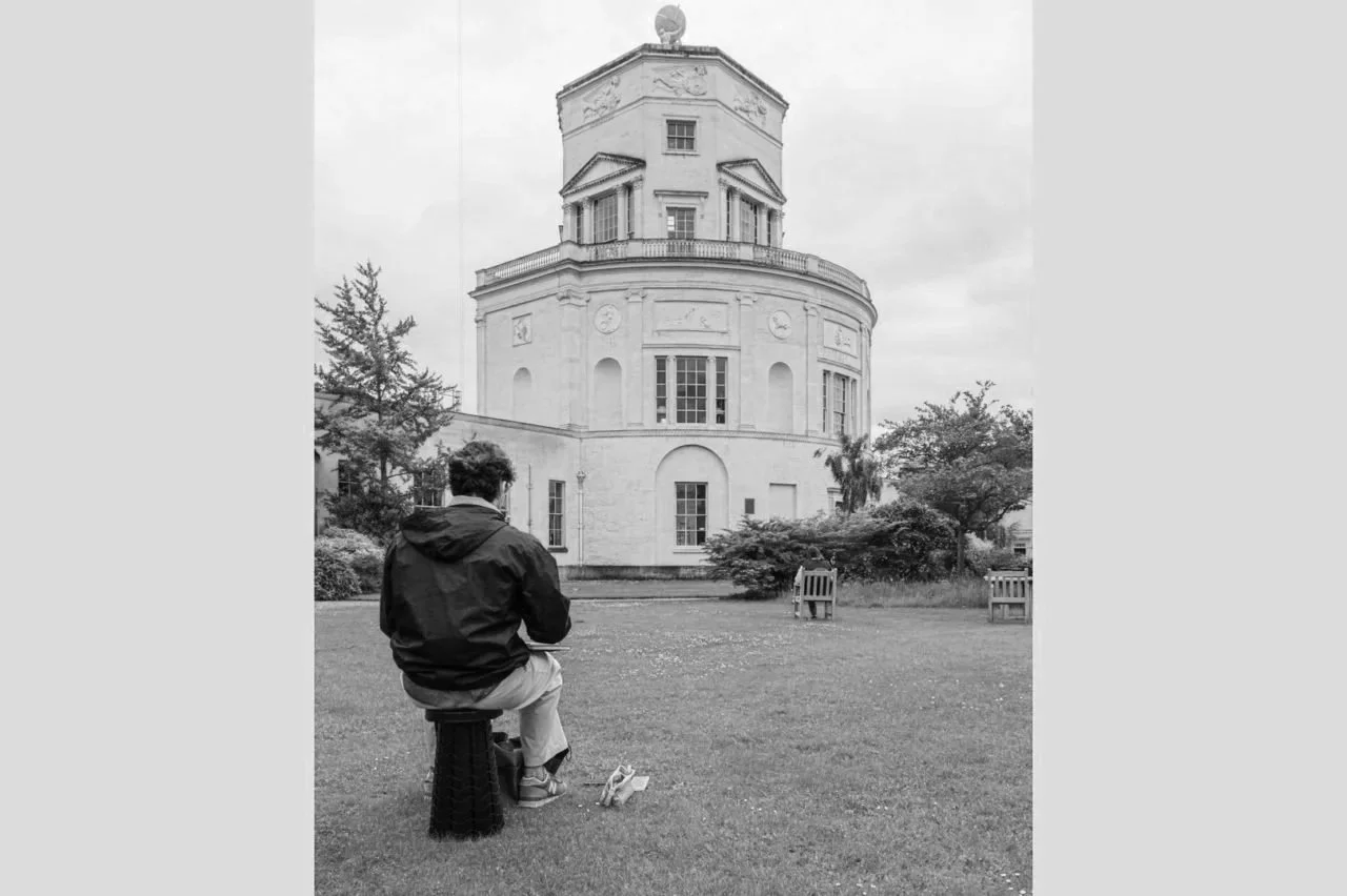 A person sitting on a stool in a park, facing a large historic building with a round tower and windows, trees, and benches nearby.