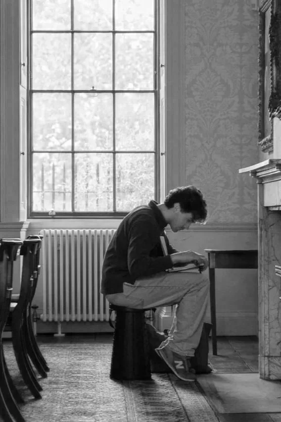 A young man sitting on a small stool by a window, reading or writing in a room with vintage decor.