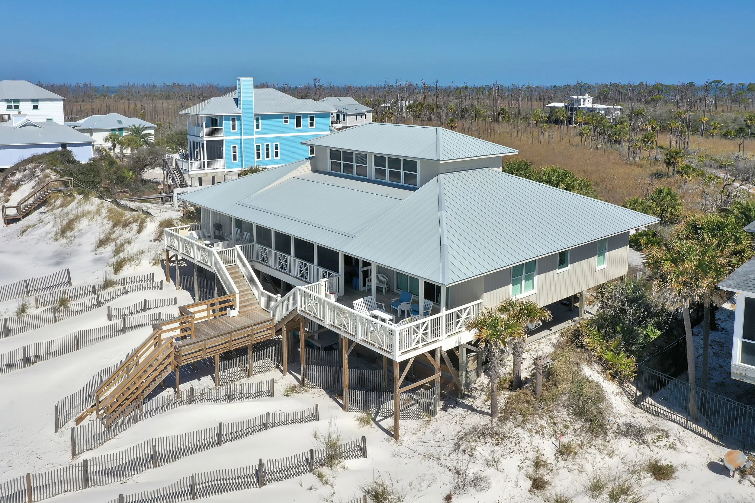 Beach house with a gray roof and white railings on stilts, surrounded by sandy dunes, palm trees, and other houses in a coastal area.