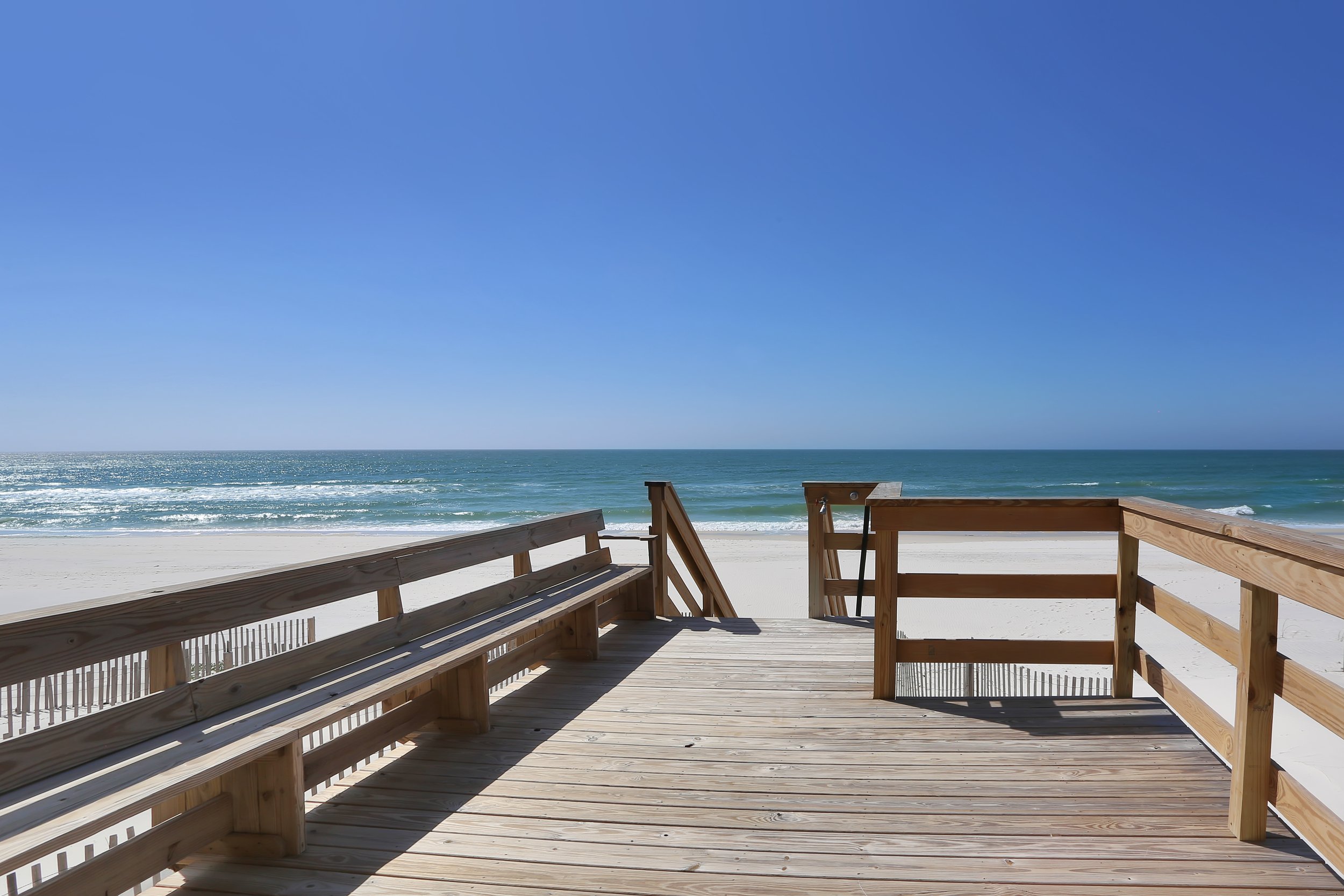 Wooden beach boardwalk leading to sandy beach and ocean with clear blue sky.