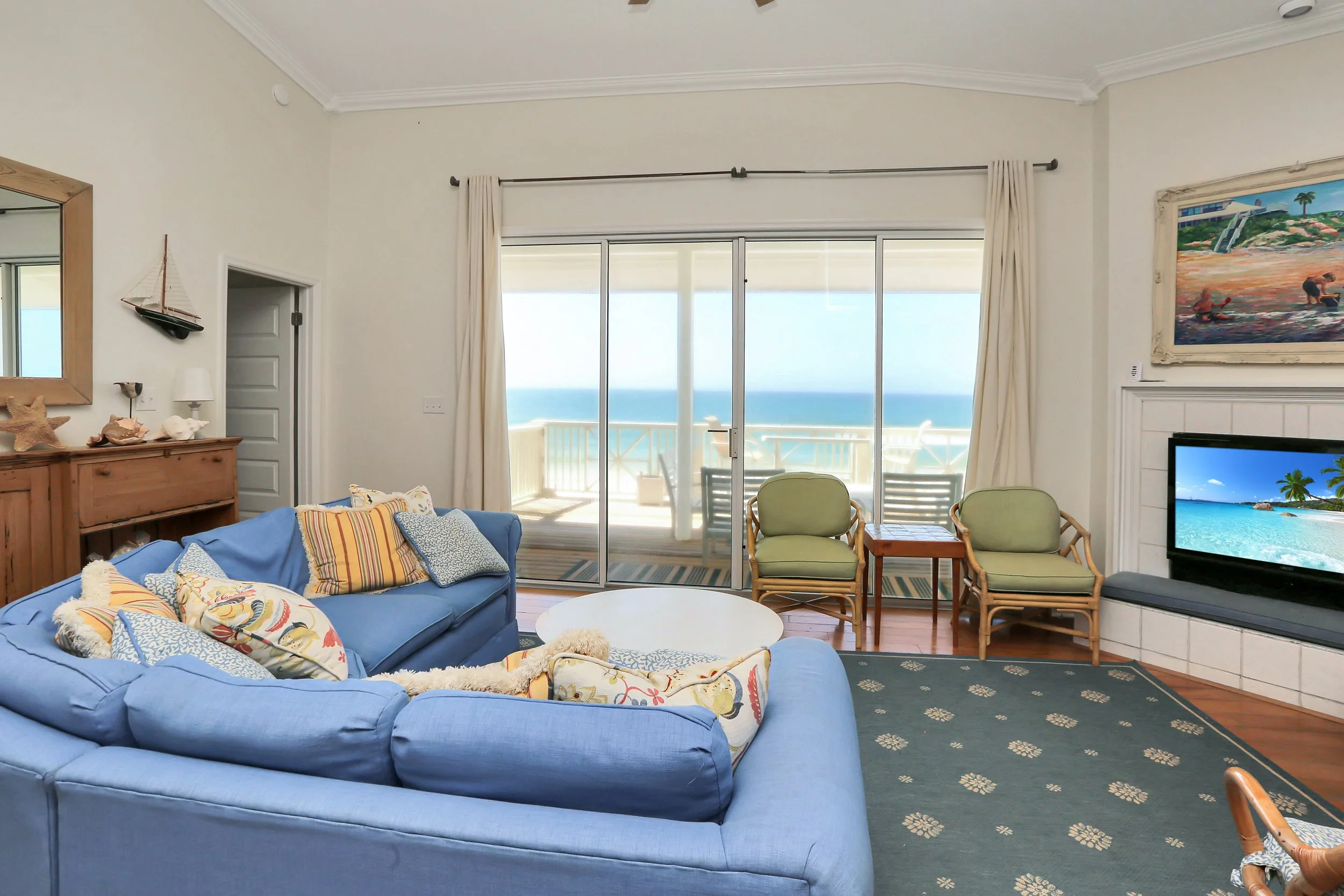 Living room with ocean view, featuring a blue sofa with patterned pillows, a wooden cabinet with decor, a flat-screen TV, and chairs by glass sliding doors leading to a balcony.