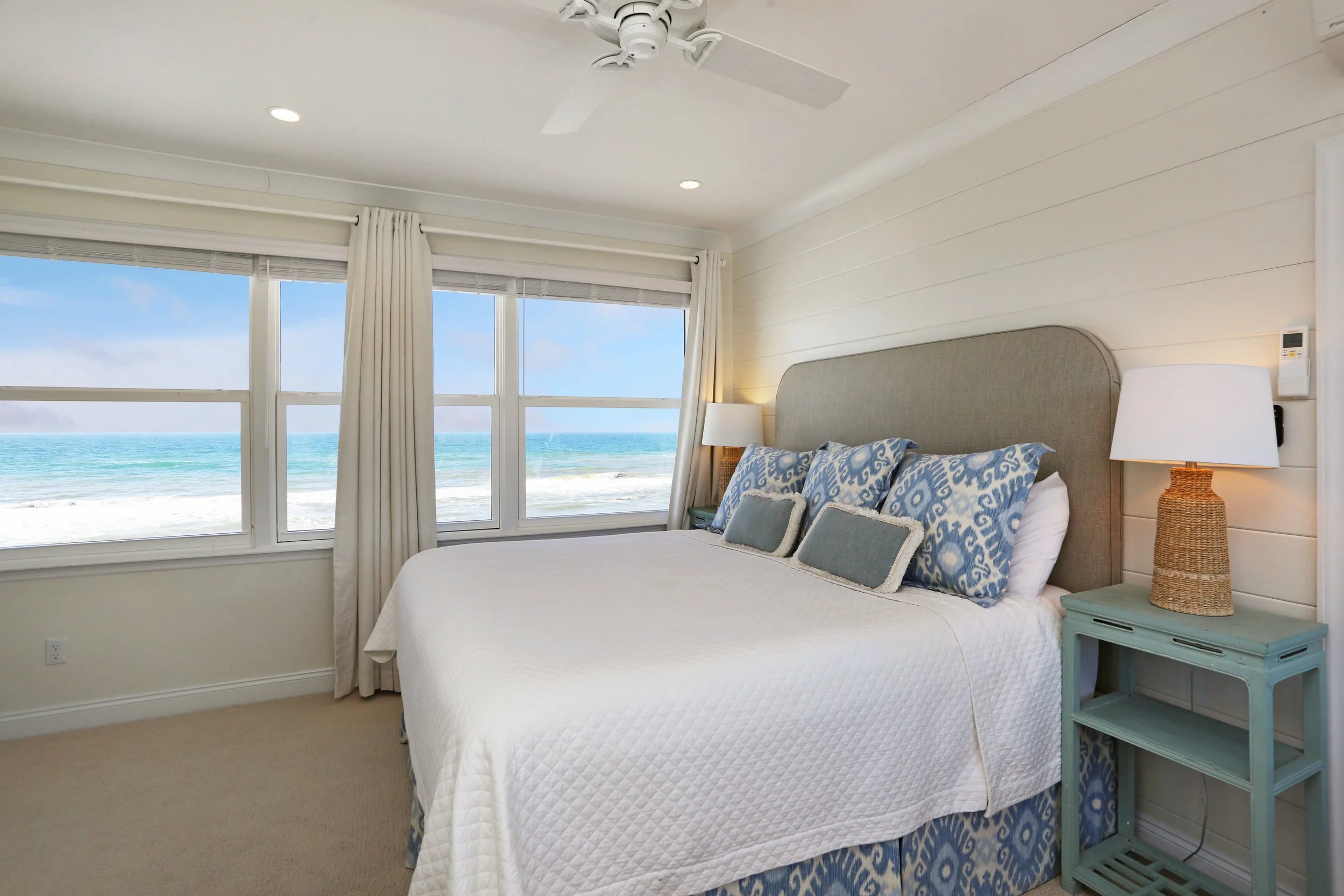 Beachside bedroom with large windows showing ocean view, furnished with a bed, decorative pillows, and bedside lamps.