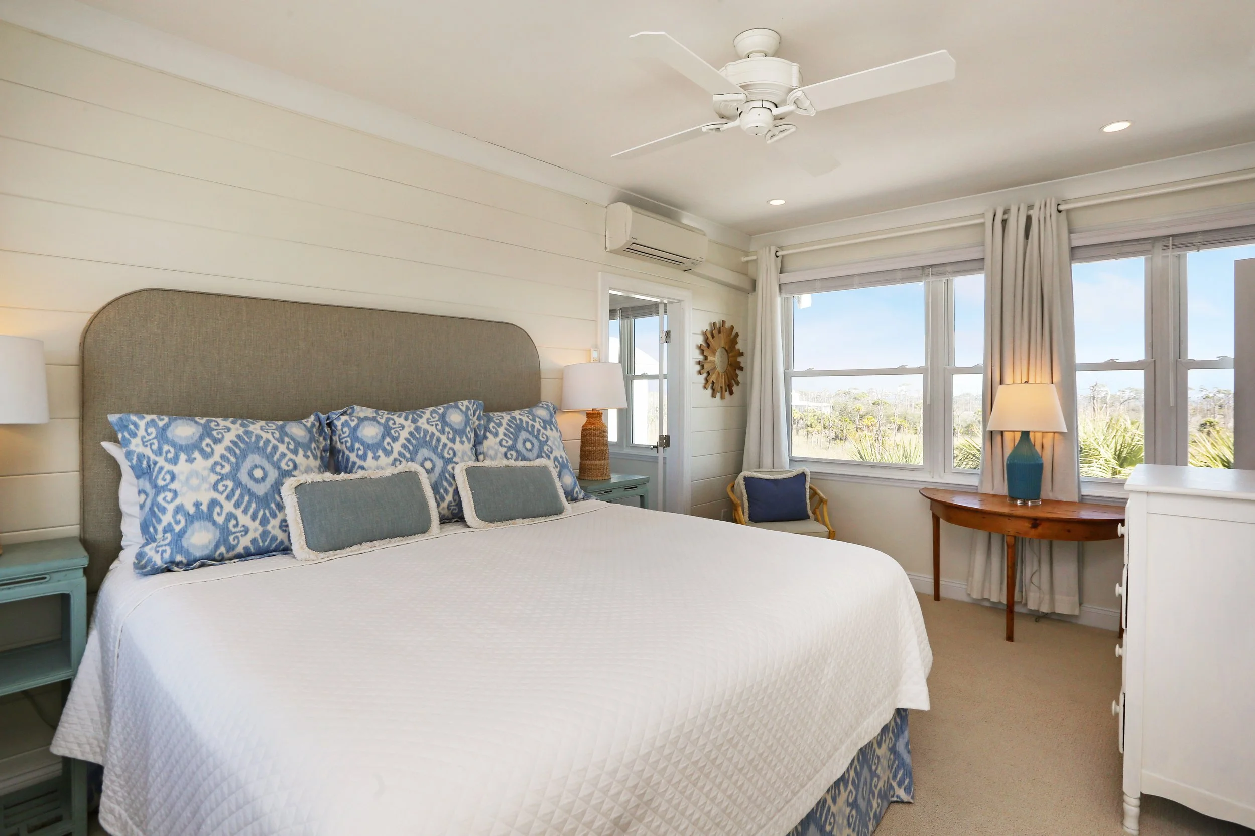 Bright bedroom with white paneled walls, large bed with white bedding and blue patterned pillows, bedside tables with lamps, window seat with blue pillows, large windows with white curtains showing an outdoor landscape, ceiling fan, and white dresser
