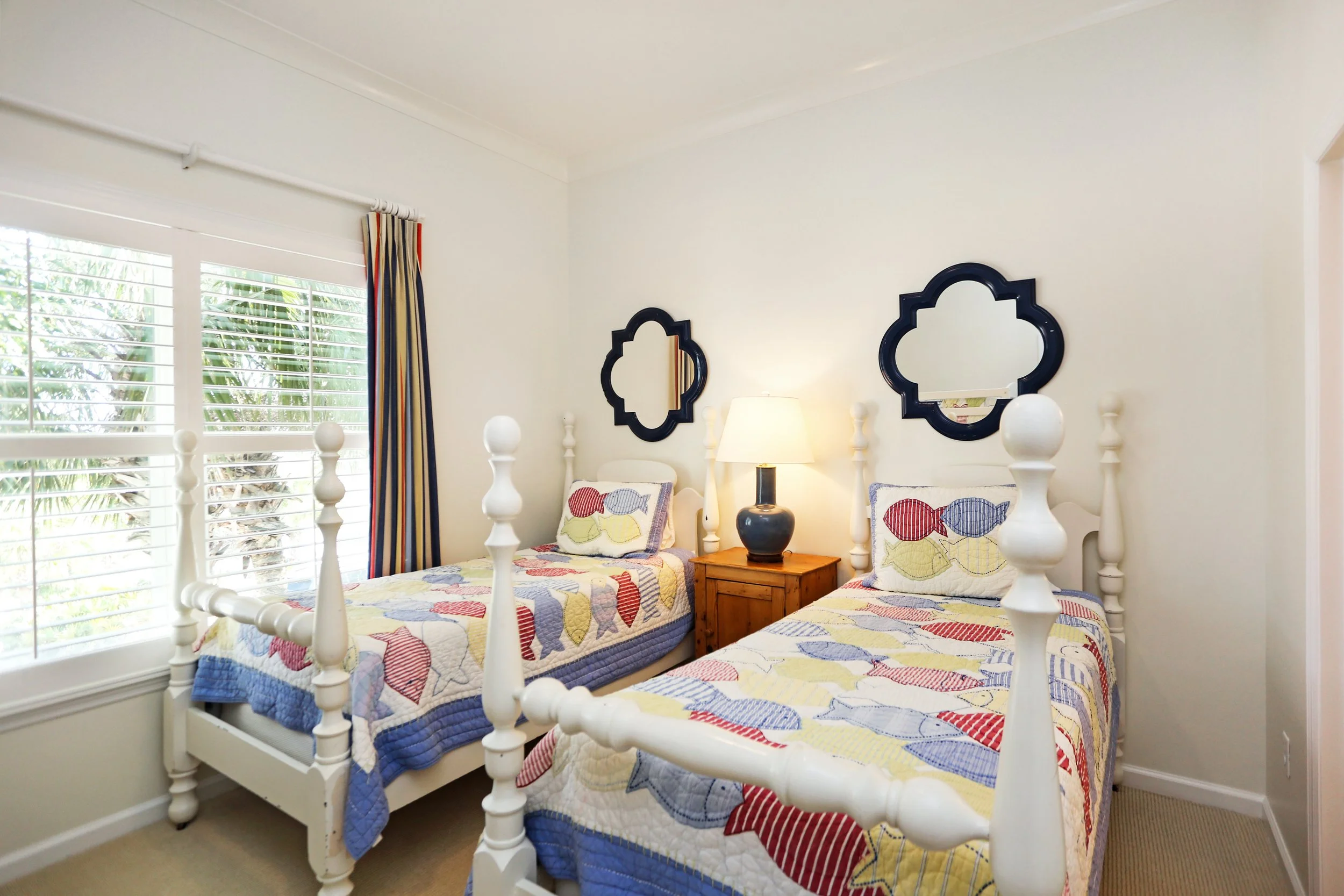 Twin bedroom with white wooden beds, quilted fish-themed bedspreads, matching pillows, bedside lamp, window with blinds and colorful curtains, and two black decorative mirrors above the beds.