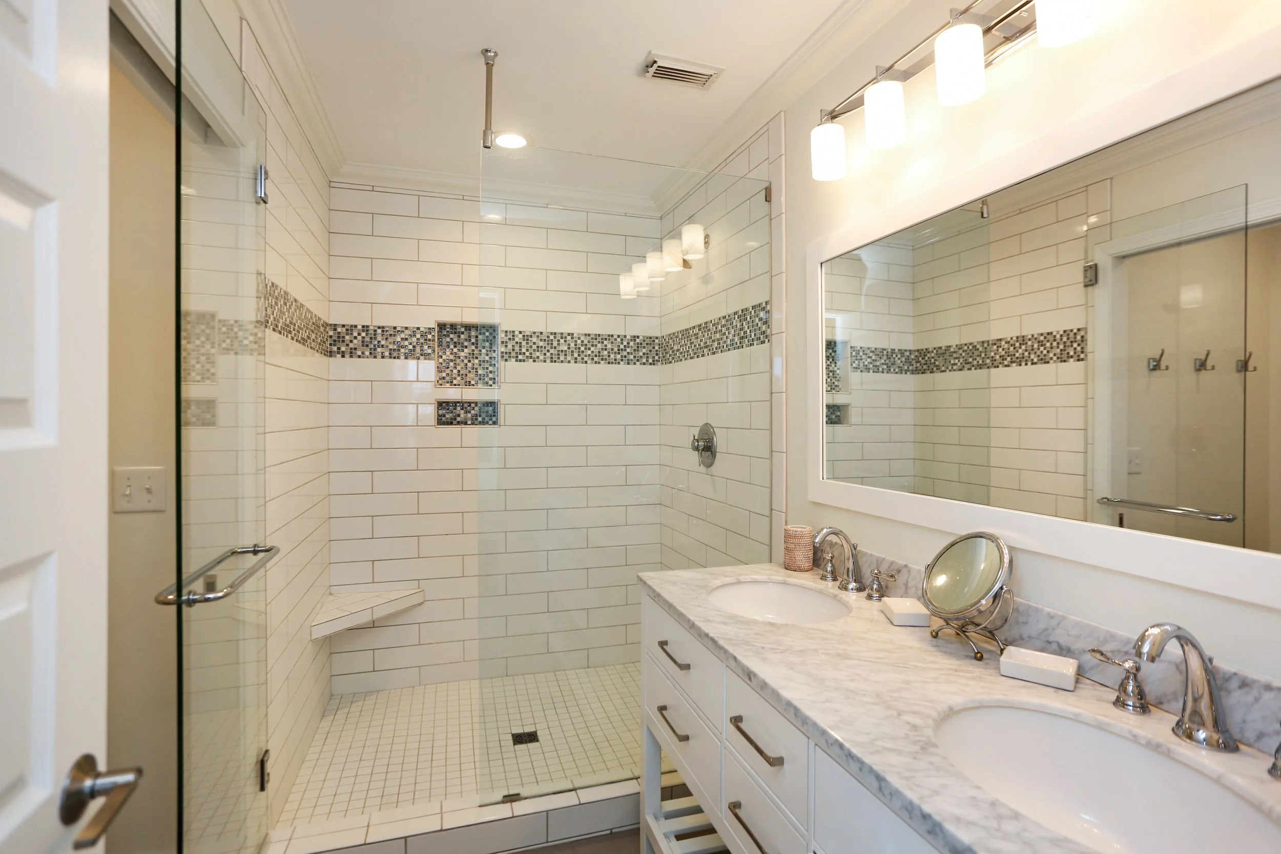 Bathroom with a walk-in shower, large mirror, marble countertop with sinks, and decorative tile accents.